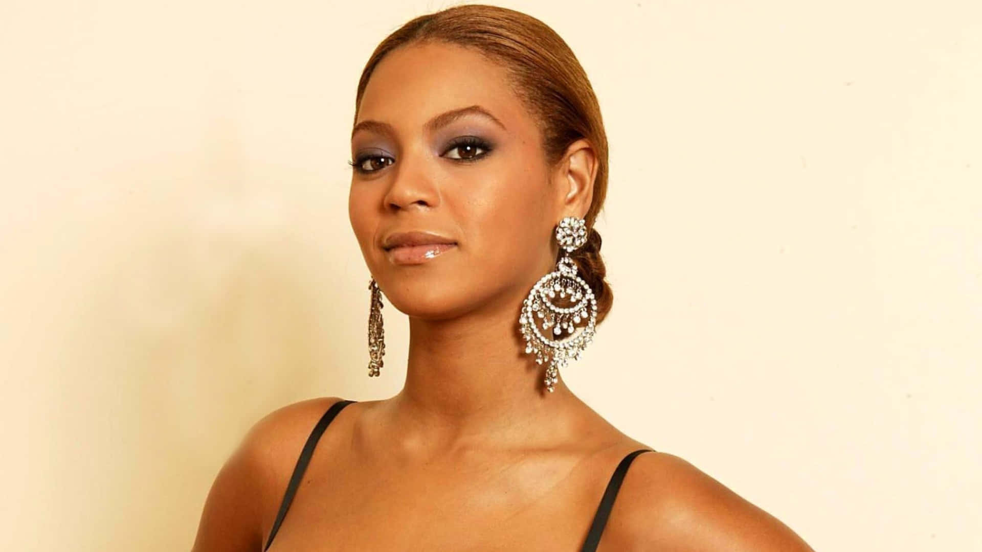 碧昂絲舞台獻唱 壁紙 — Beyoncé 自信地擺姿勢，身穿黑色上衣，佩戴精緻的鑽石耳環，背景是柔和的米色。