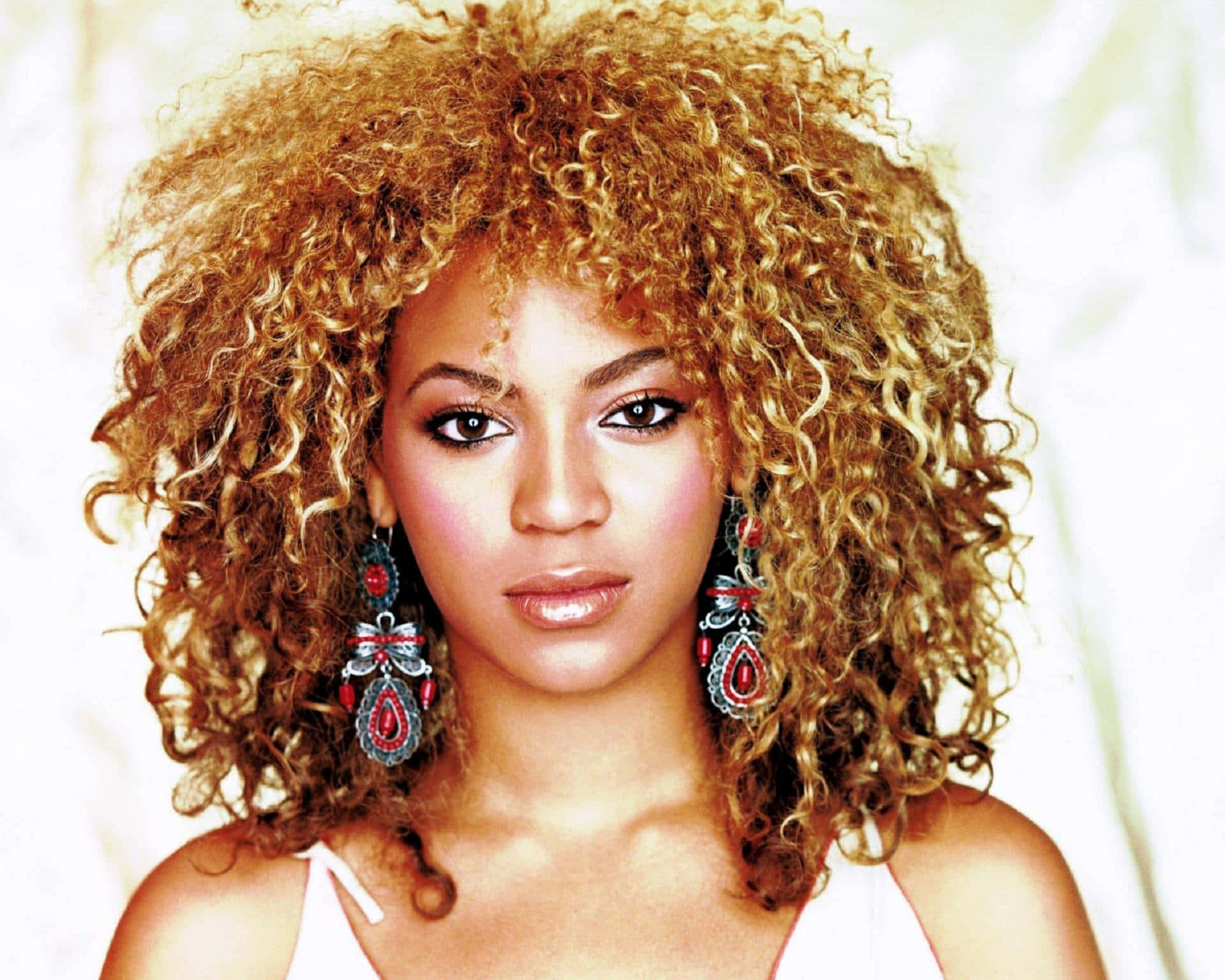 碧昂絲身披金光璀璨 壁紙 — Beyoncé 擁有豐盈金色捲髮和華麗紅色耳環，直視柔和的白色背景。