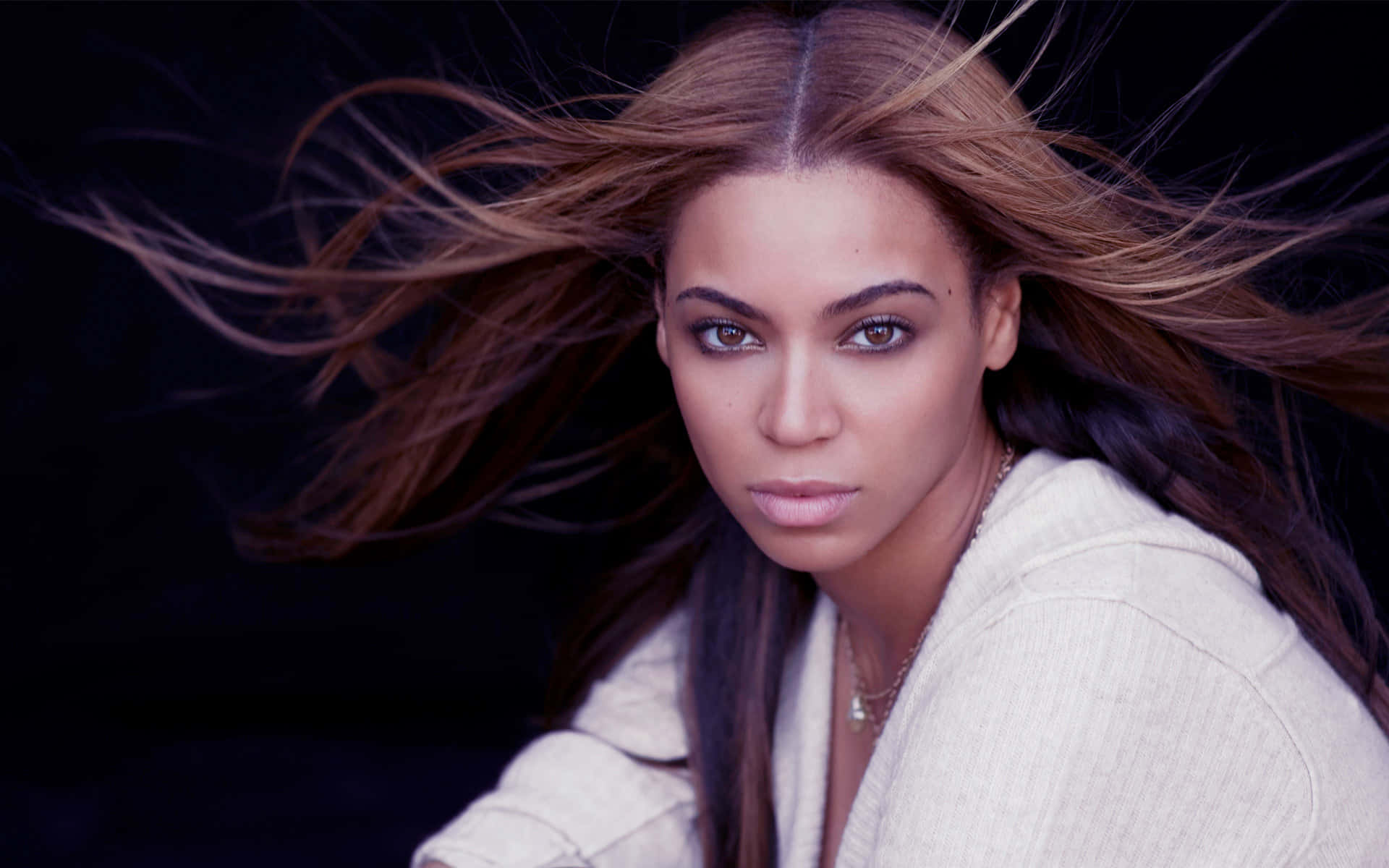碧昂絲身著華麗金裙挺拔而立 壁紙 — Beyoncé 在深色背景下，棕色頭髮隨風飄揚，穿著白色連帽衫，凝視著。