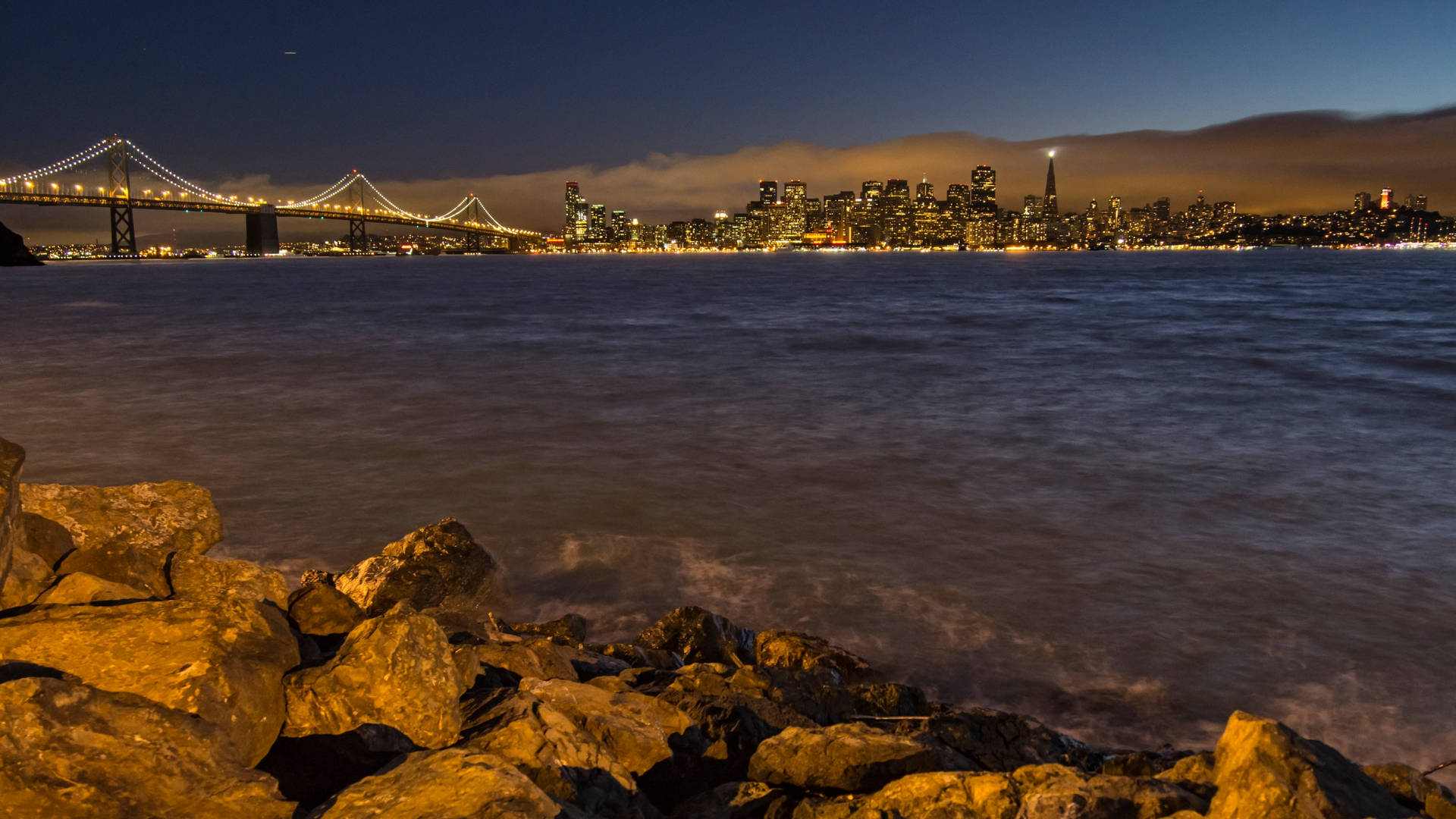 從岩石海岸觀看三藩市天際線和海灣大橋的夜景。