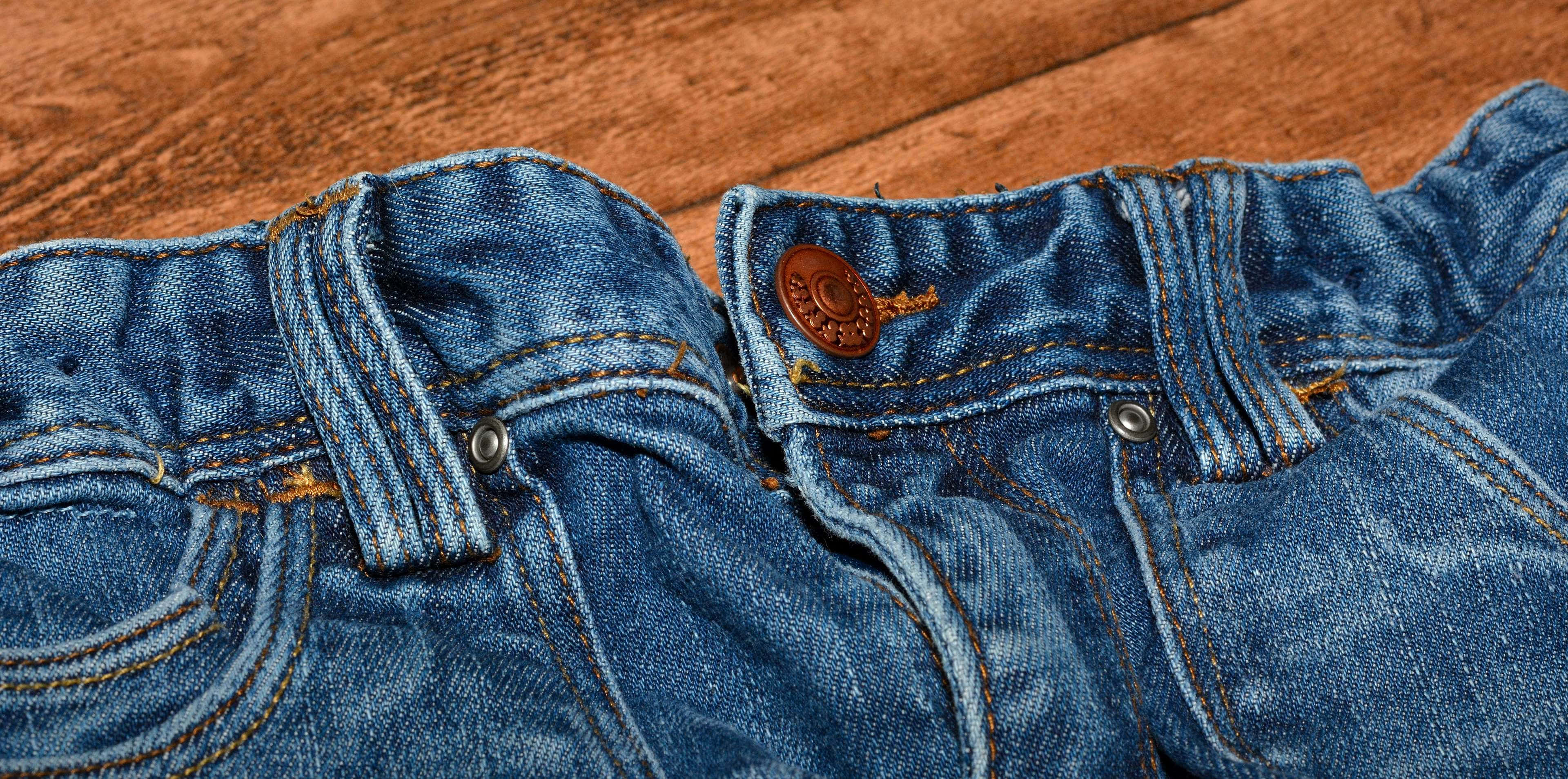 Denim 桌布 壁紙 — 磨損嘅藍色牛仔褲特寫，有啡色縫線同鈕扣，放喺溫暖嘅木質表面上。