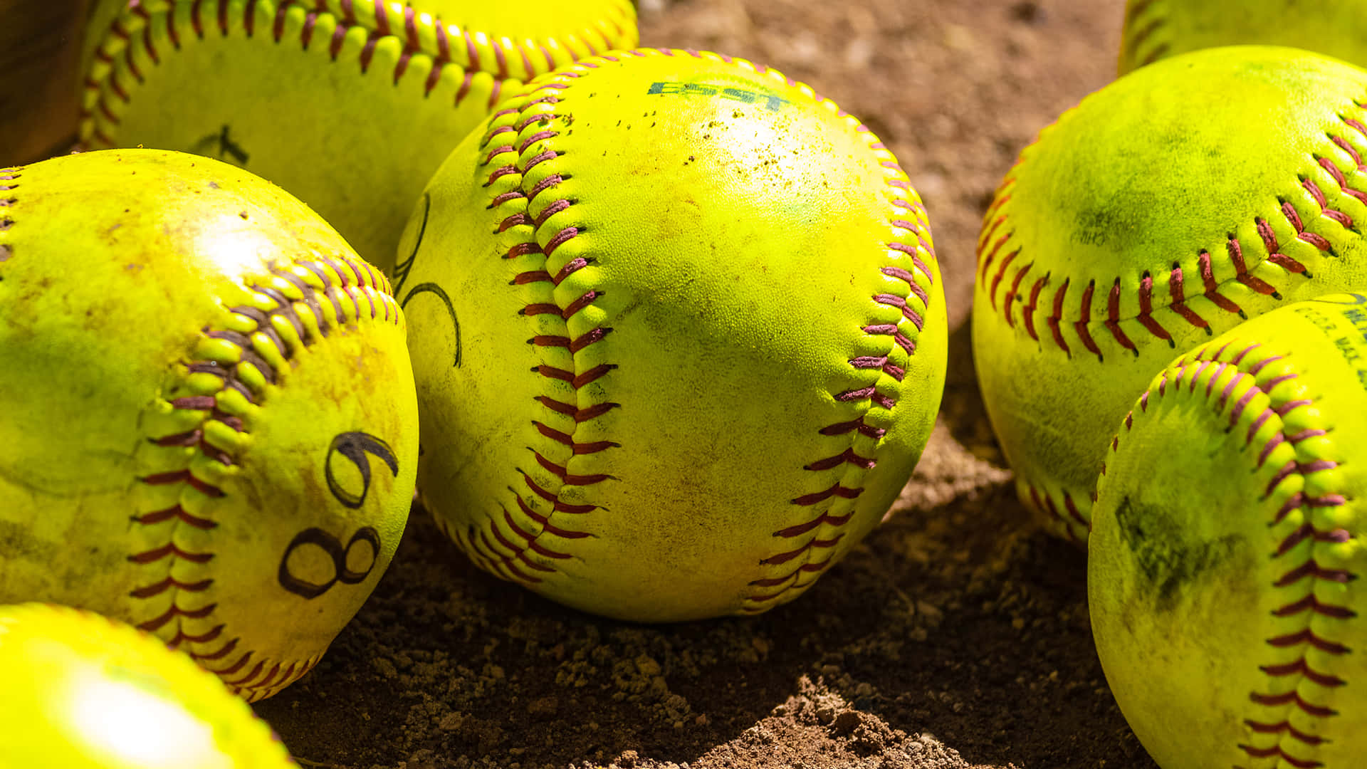 Softball 桌布 壁紙 — 磨損的黃色壘球特寫，帶有紅色縫線，放置在棕色泥土場地上。