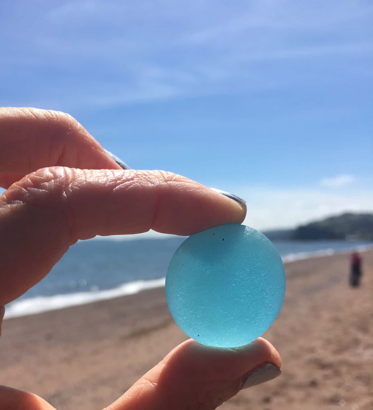 磨砂海玻璃圓形圖 壁紙 — 一隻手拿著一個光滑半透明的藍色海玻璃球體，背景是陽光明媚的海灘，有輕柔的海浪和遠處的山丘。
