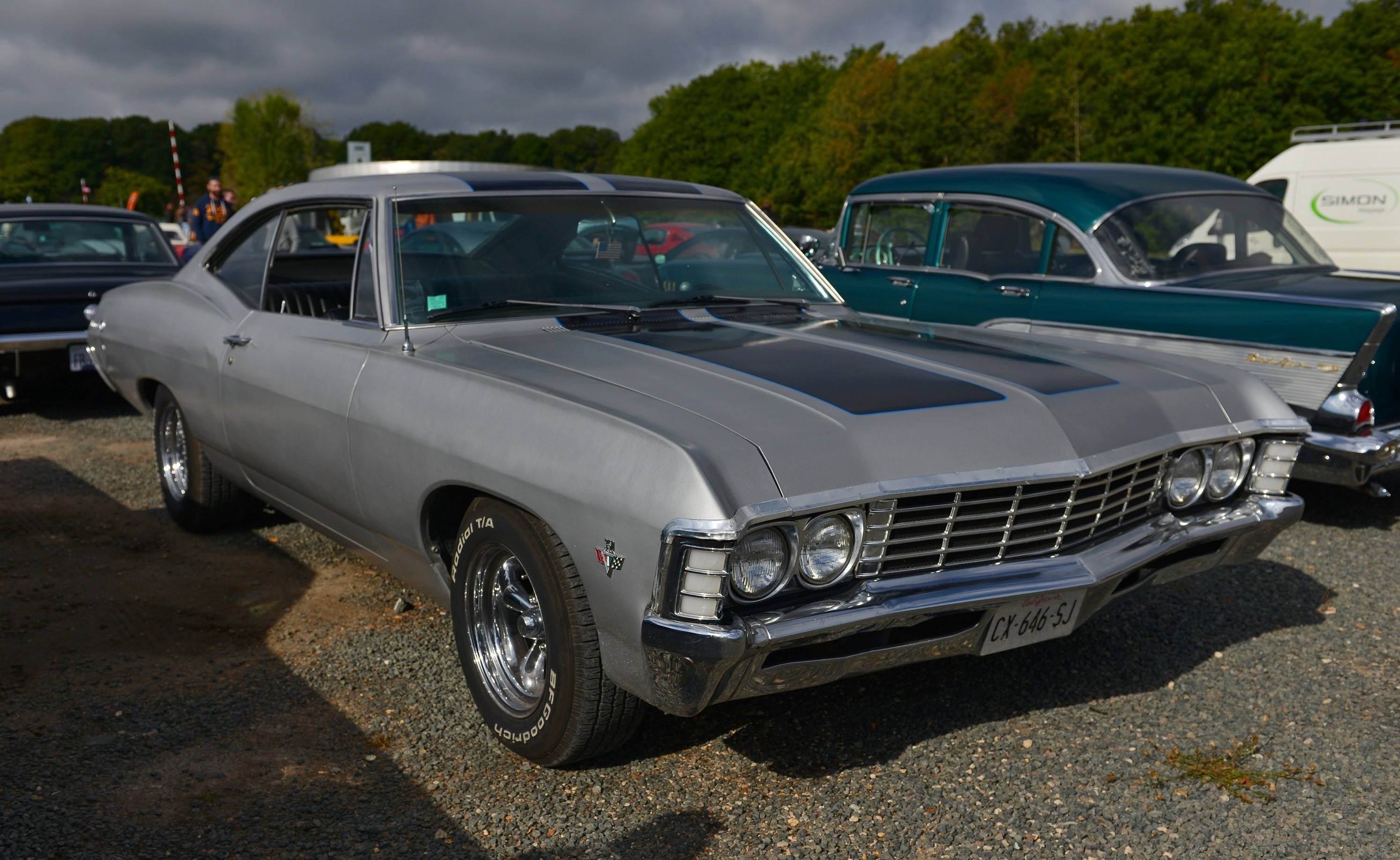 磨砂灰雪佛蘭Impala 1967 壁紙 — 一輛閃亮的銀色經典肌肉車，帶有黑色引擎蓋條紋，停在碎石地上，在陰天下的車展中。