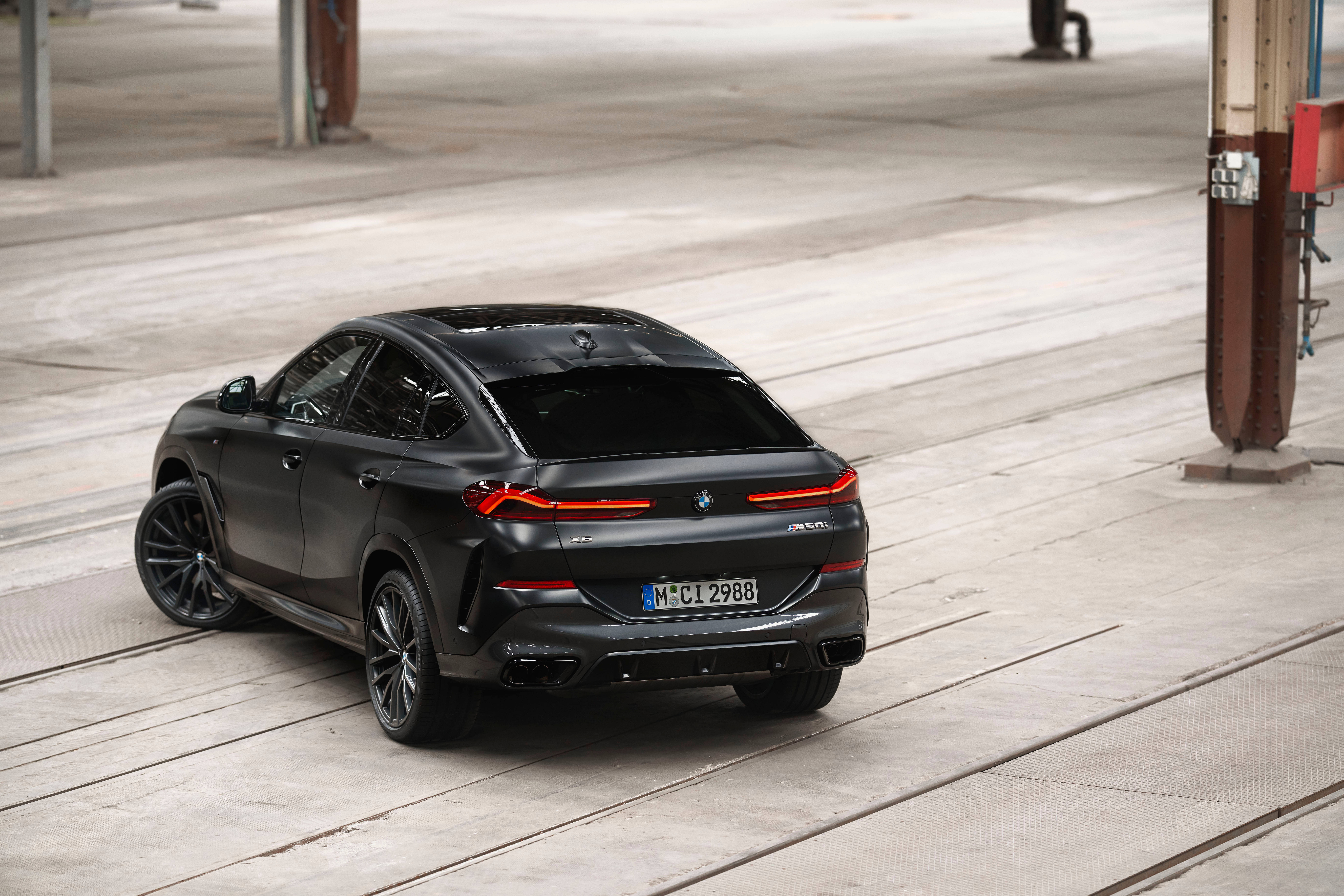 磨砂黑 BMW X6 壁紙 — 一輛啞光黑色BMW X6 M停泊在工業橫樑下的混凝土軌道上，紅色尾燈發光。