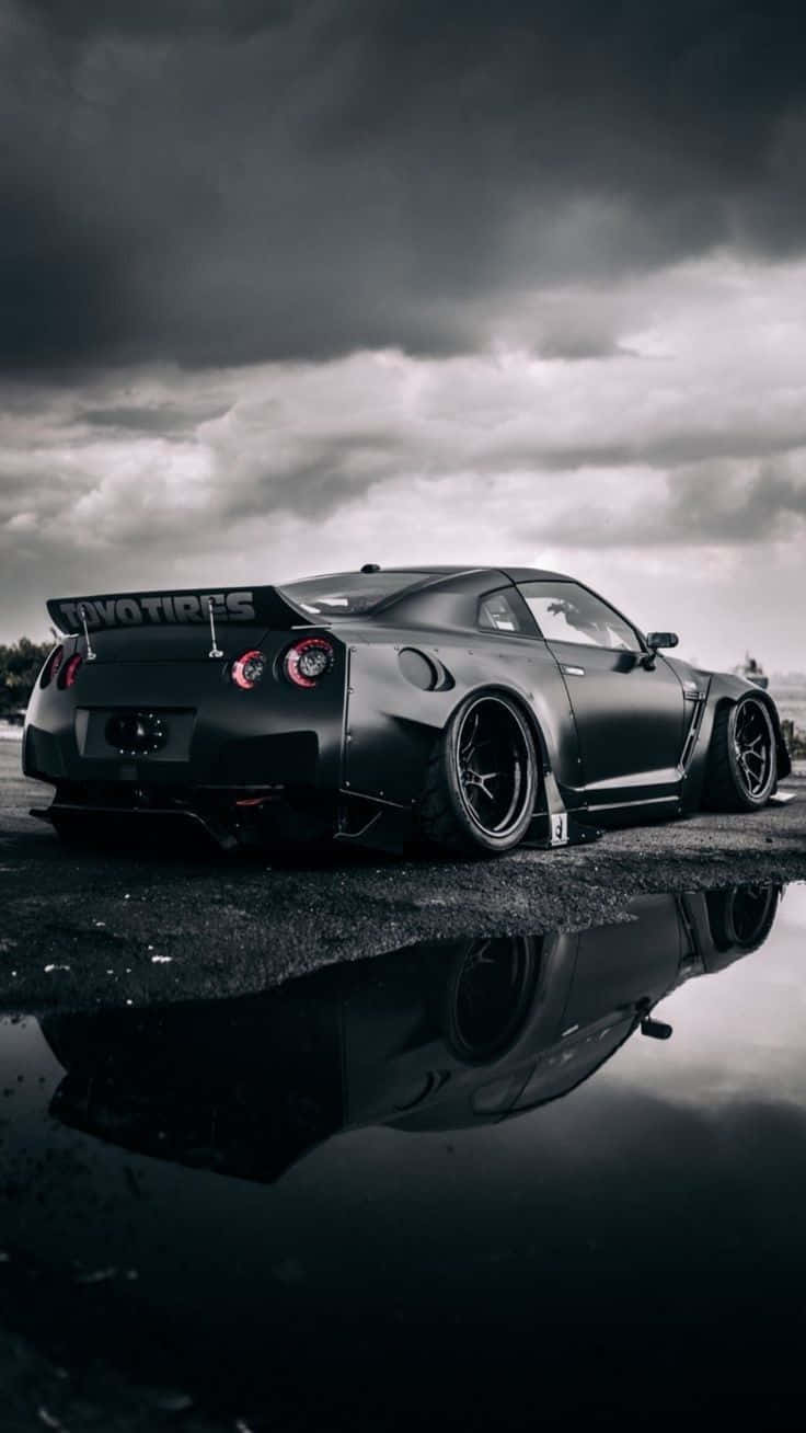 磨砂黑 Nissan R35 GT-R 水面倒影圖 壁紙 — 一輛啞光黑色的日產GT-R配備寬體套件，停在暴風雲下的濕瀝青路面上，倒影映在水坑中。