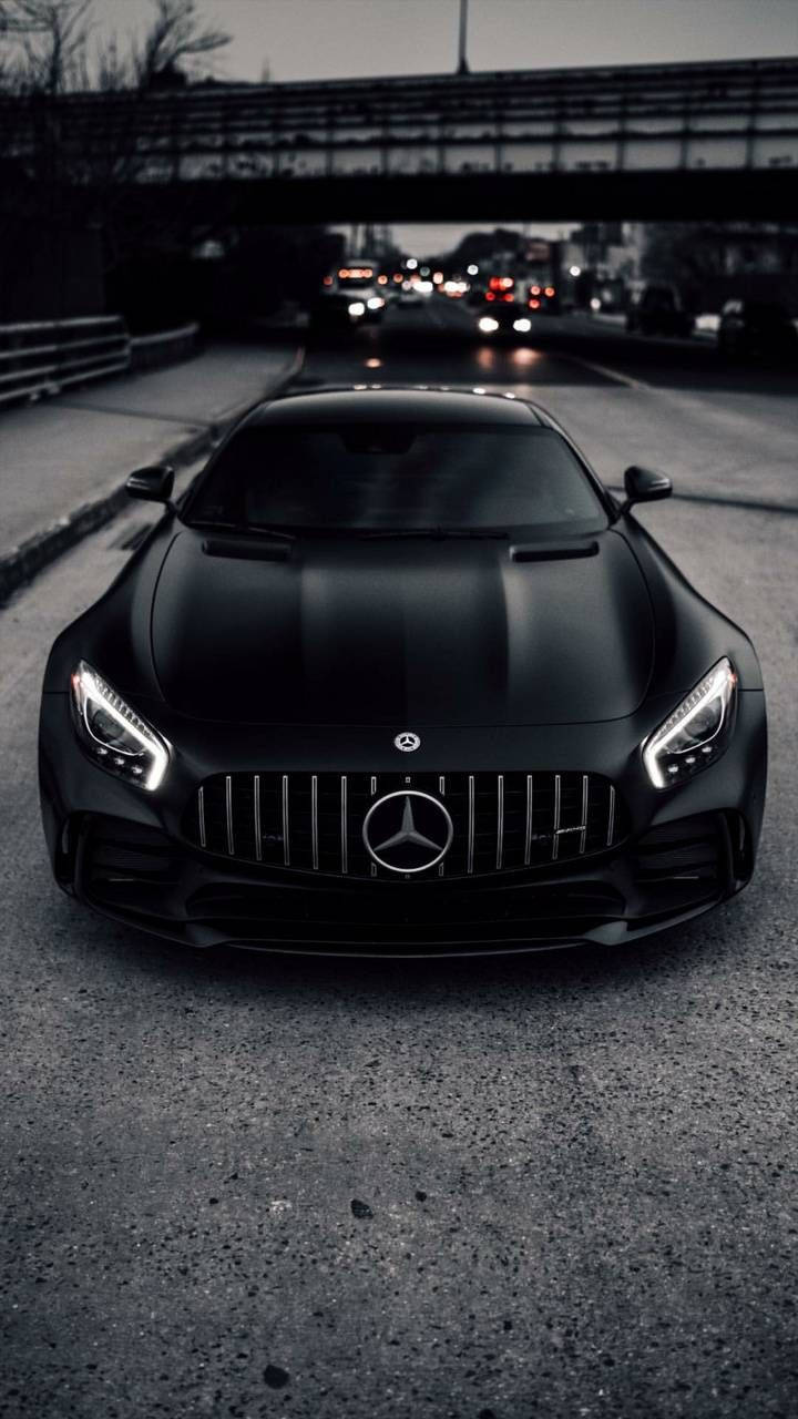 磨砂黑AMG行駛於道路 壁紙 — 一輛啞光黑色的 Mercedes-AMG GT 跑車在橋下昏暗的城市道路上向前行駛，遠處模糊的交通燈閃爍著光芒。