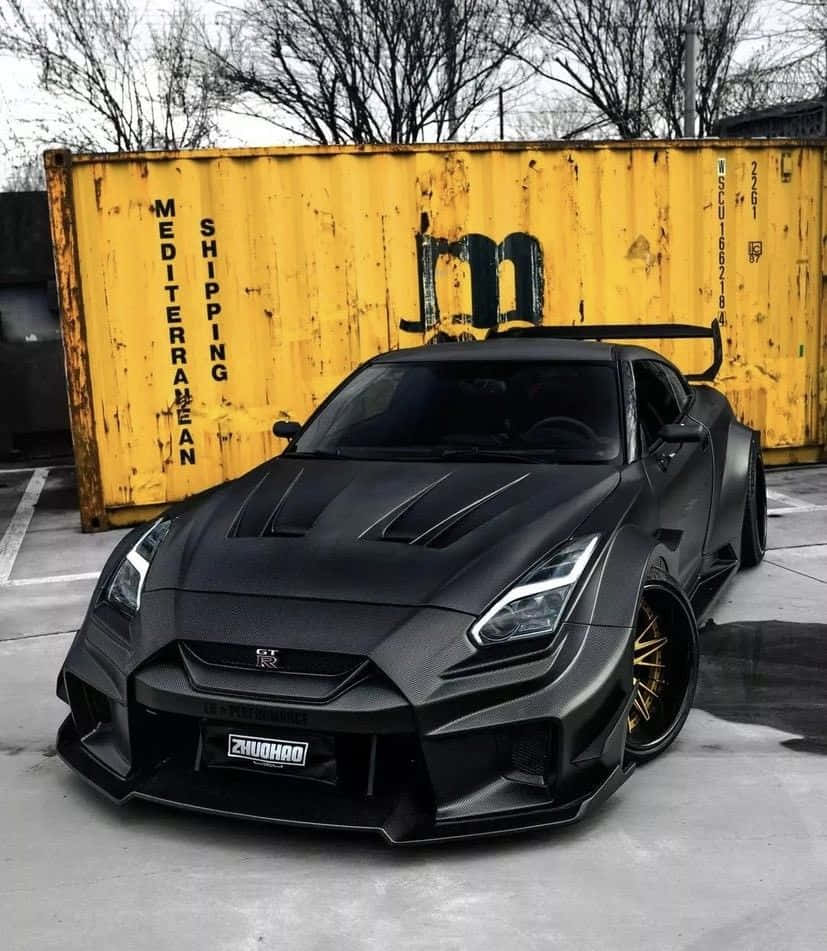 磨砂黑Nissan R35 GT-R圖 壁紙 — 一架啞黑色改裝日產GT-R跑車，配金色輪圈，停泊在一個風化了的黃色貨櫃前面。