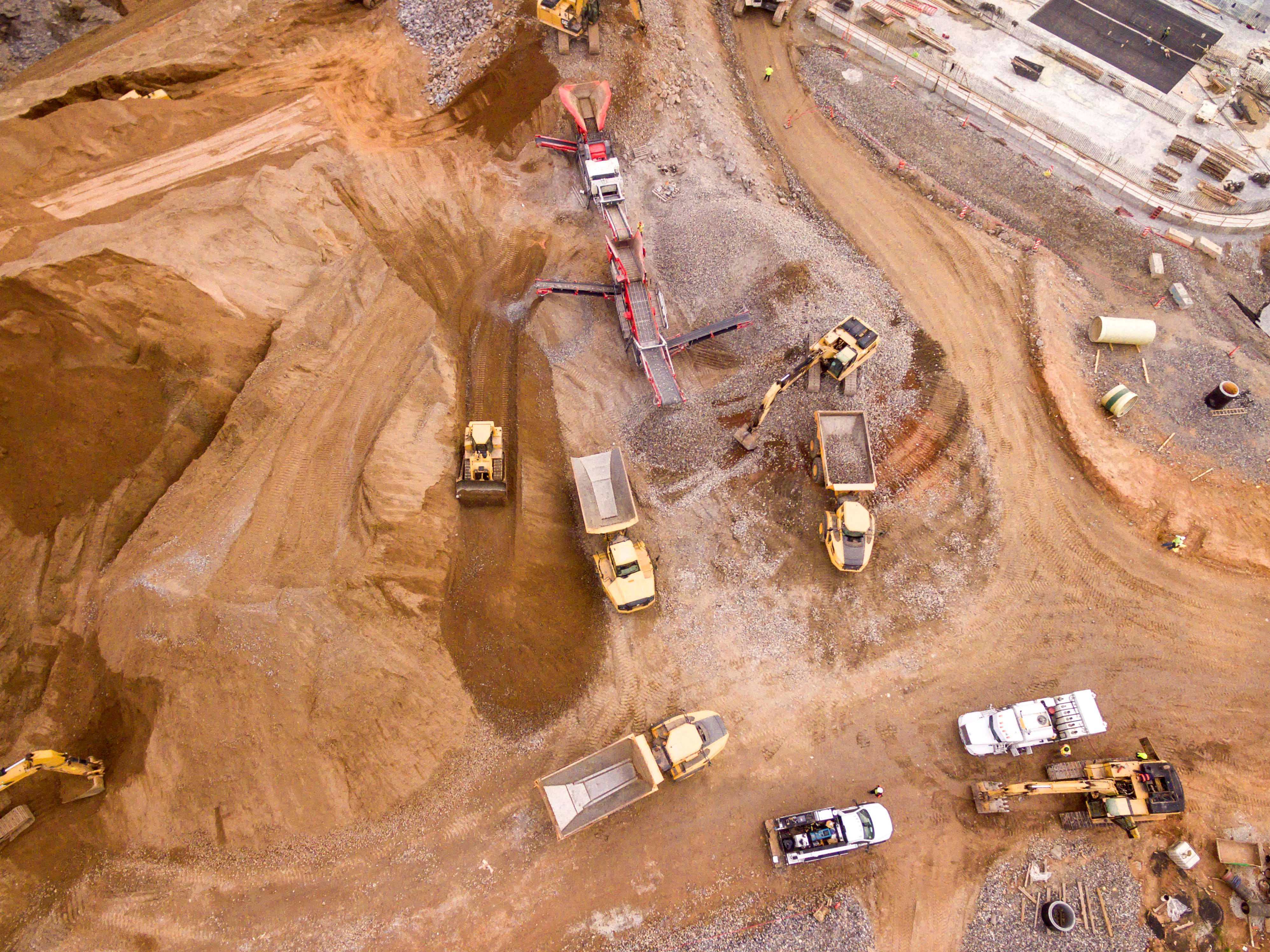 礦場卡車 壁紙 — 繁忙建築工地的鳥瞰圖，重型機械在明亮的日光下移動泥土和礫石。