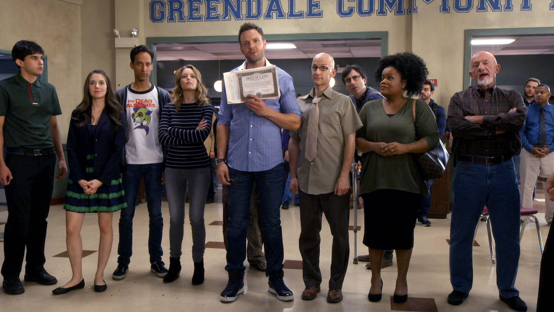 社區畢業證書 壁紙 — 一群多元化的成年人站在學校走廊，上方有「Greendale Community」標誌，一名男子手持裱框證書，其他人則在觀看。