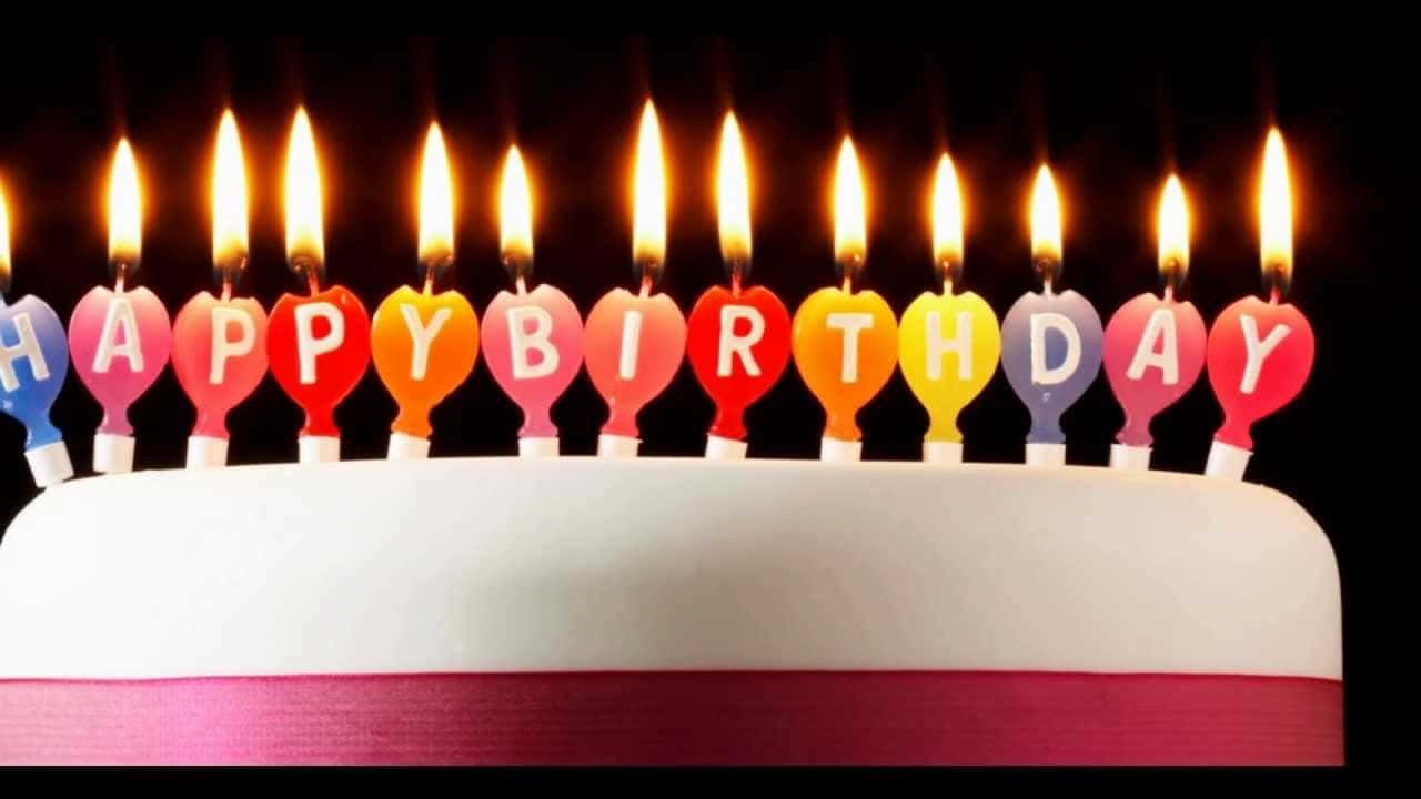 祝你度過一個美麗難忘的生日！ 壁紙 — 一個白色生日蛋糕，上面裝飾著彩色心形蠟燭，拼出「HAPPY BIRTHDAY」，全部點燃，背景為黑色。
