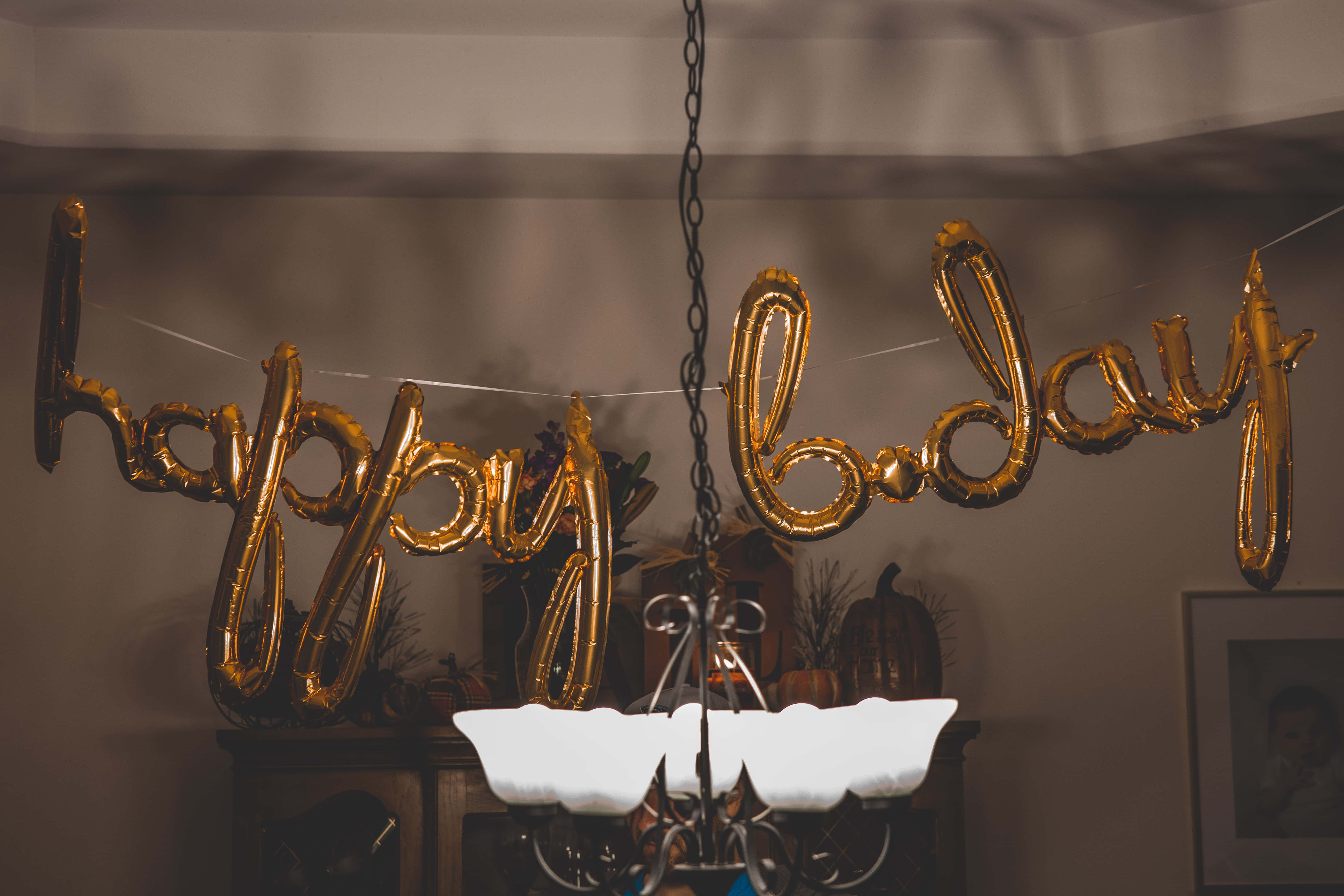 祝你生日快樂！ 壁紙 — 金色 'happy birthday' 氣球懸掛在點亮的吊燈上方，營造出溫暖的室內慶祝場景。