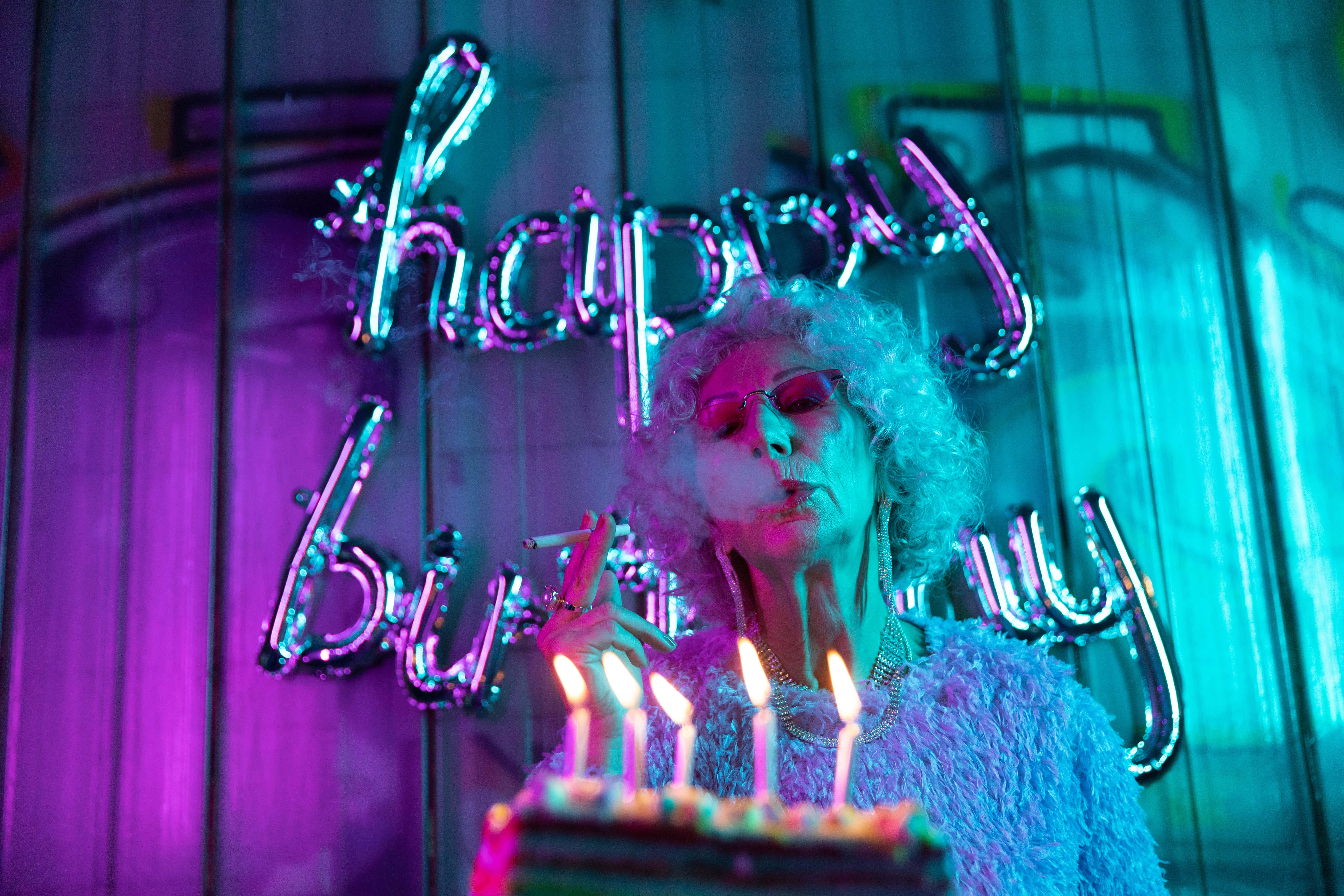 祝你生日快樂，充滿光明與喜悅！ 壁紙 — 一位擁有捲髮和太陽眼鏡的女性，在發光的 'happy birthday' 霓虹燈牌下吹熄生日蛋糕上的蠟燭。