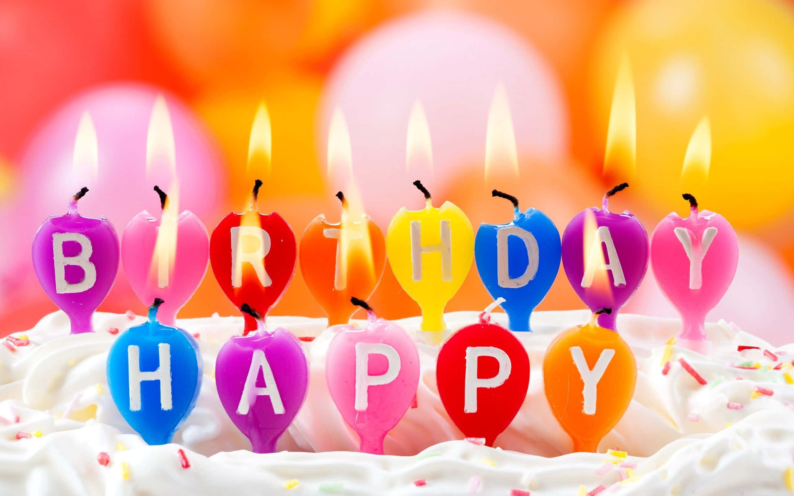 祝你生日快樂且福氣滿滿！ 壁紙 — 充滿活力的心形生日蠟燭拼出「HAPPY BIRTHDAY」，在鋪滿彩色糖霜的蛋糕上閃耀，背景是模糊的氣球。