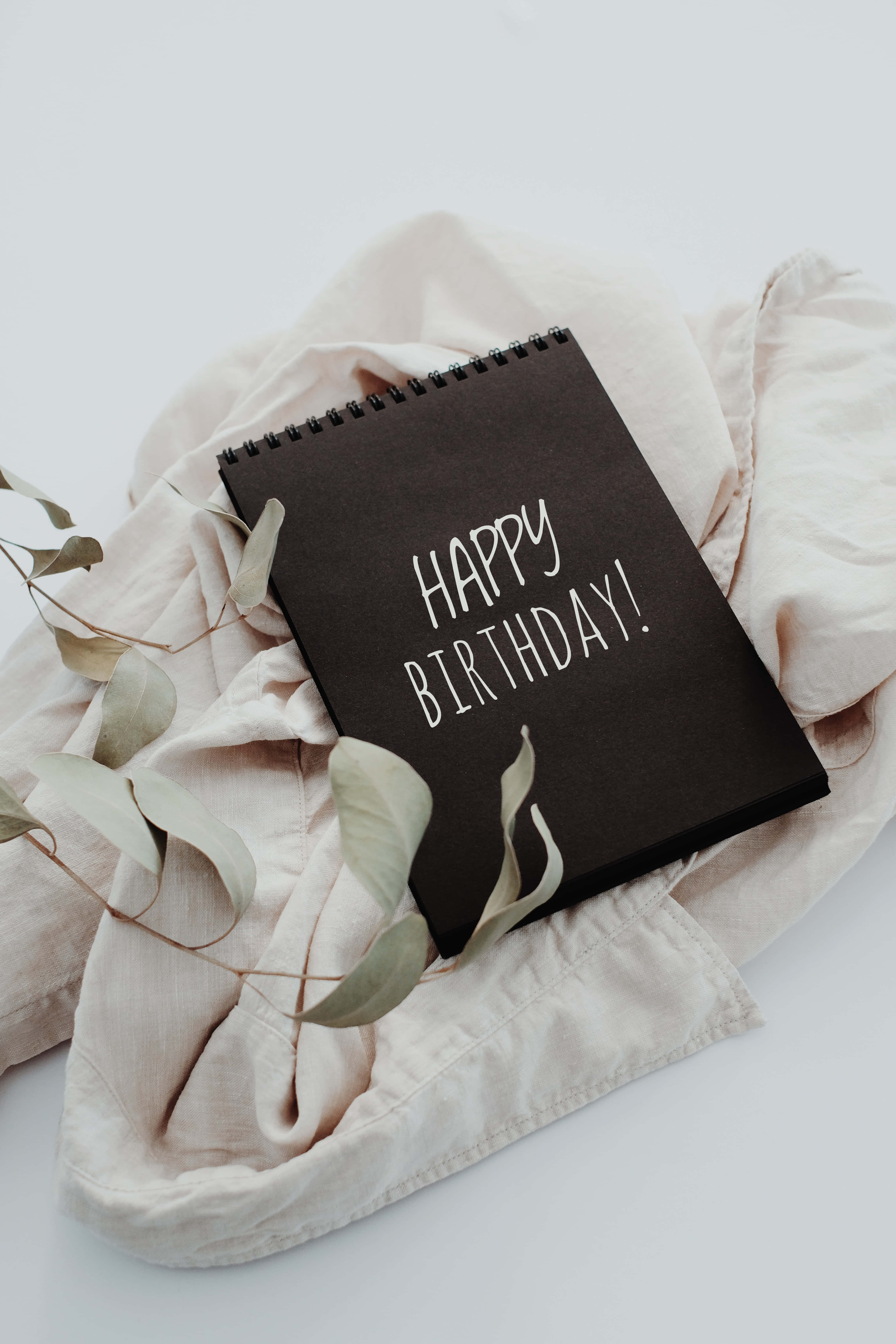 祝你生日最快樂！ 壁紙 — 一本黑色螺旋筆記本，上面有白色手寫字體的「HAPPY BIRTHDAY!」，放在柔軟的米色布料上，搭配乾燥的尤加利葉。
