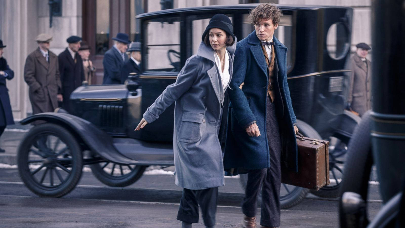 一男一女穿著1920年代服裝，在積雪街道上走過一輛復古汽車。