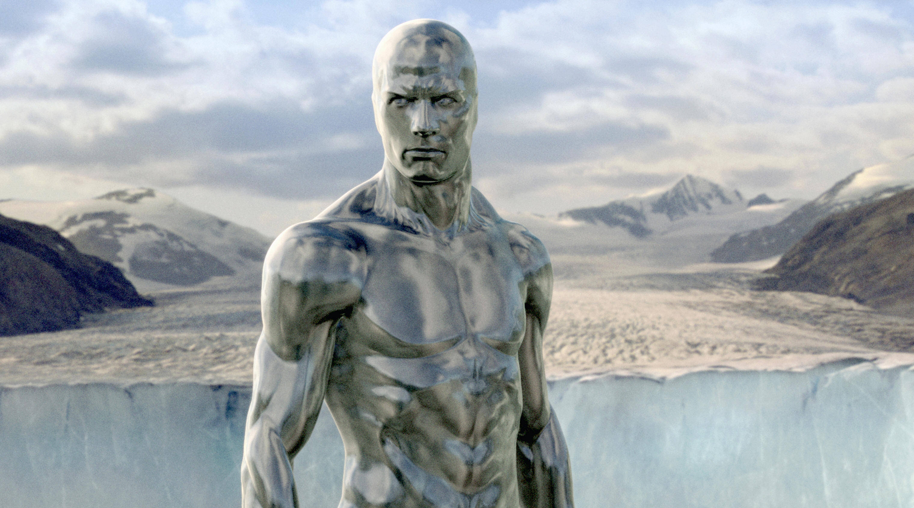 神奇四俠銀河騎士山景 壁紙 — 一個金屬銀色的肌肉男雕像矗立在廣闊的冰川前，背景是白雪覆蓋的山脈和陰天。