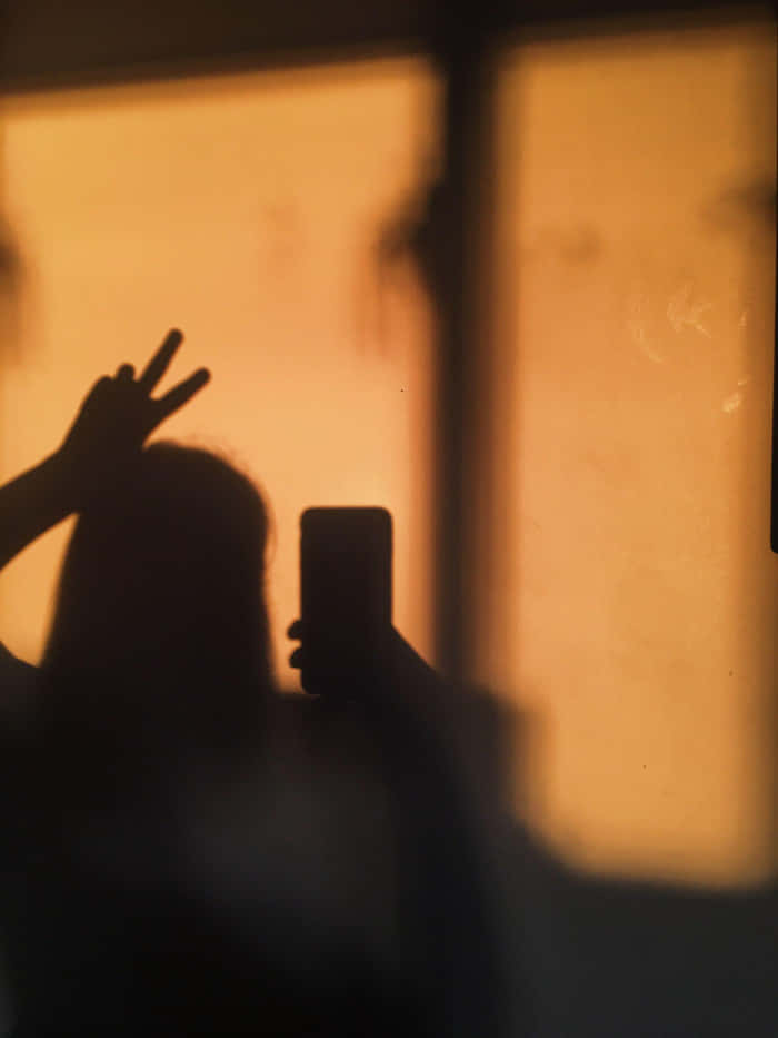 神秘剪影中的女孩側影 壁紙 — 一個人的剪影，手持電話並在溫暖發光的窗戶前做出和平手勢。