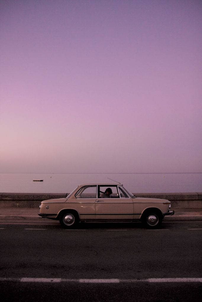 一輛米色復古車在黃昏時沿著海邊道路行駛。