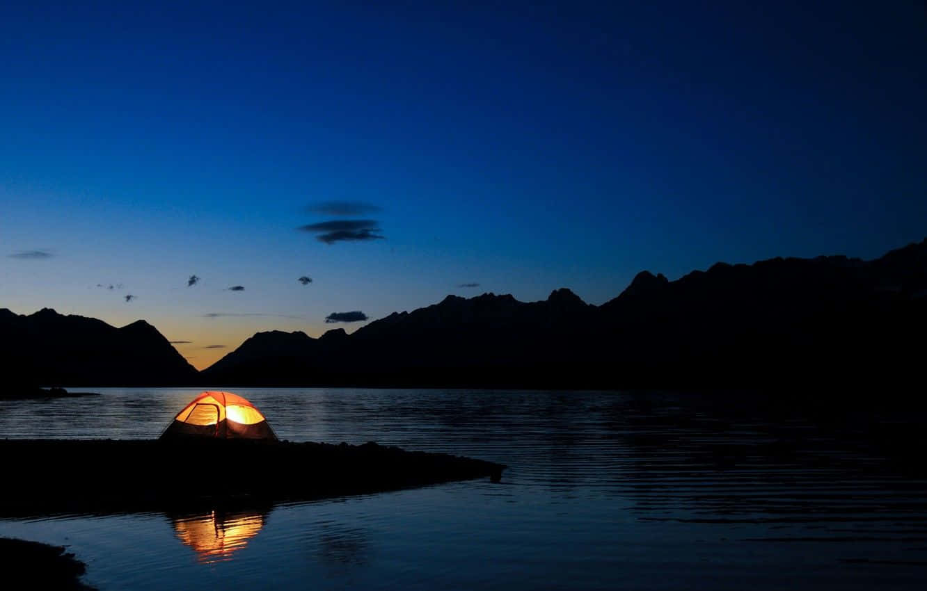 神秘黑暗風景 壁紙 — 一個發光的橙色帳篷獨自矗立在暮色中的湖岸上，其溫暖的光芒映照在平靜的水面上，與黑暗的山巒輪廓形成對比。
