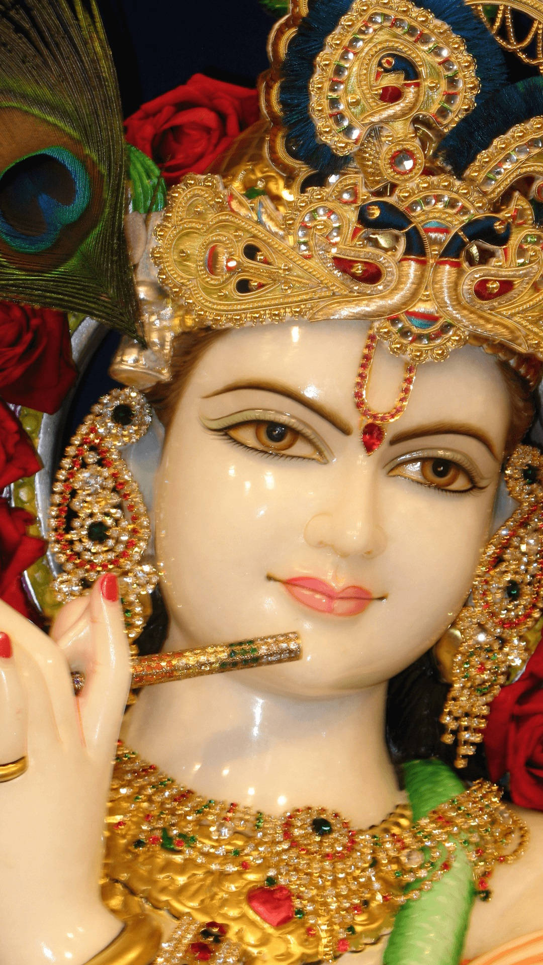 krishna iPhone 桌布 壁紙 — 神聖雕像的特寫，飾有黃金首飾、孔雀羽毛和紅玫瑰。