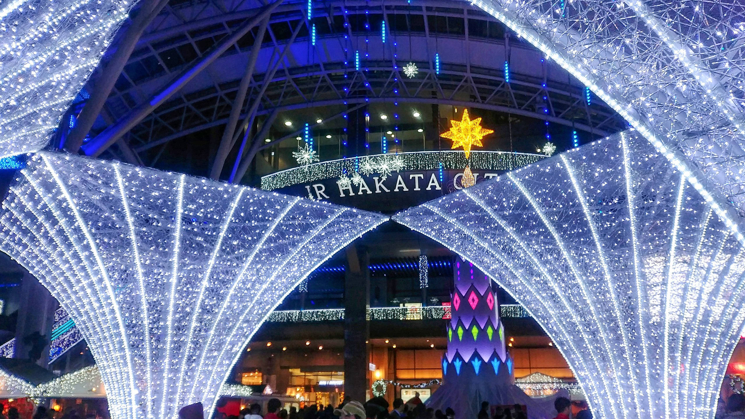 福岡 Hakata 站夕照景觀 壁紙 — 一個耀眼的藍白燈光裝置拱形橫跨在繁忙的車站入口，裝飾著節日裝飾。