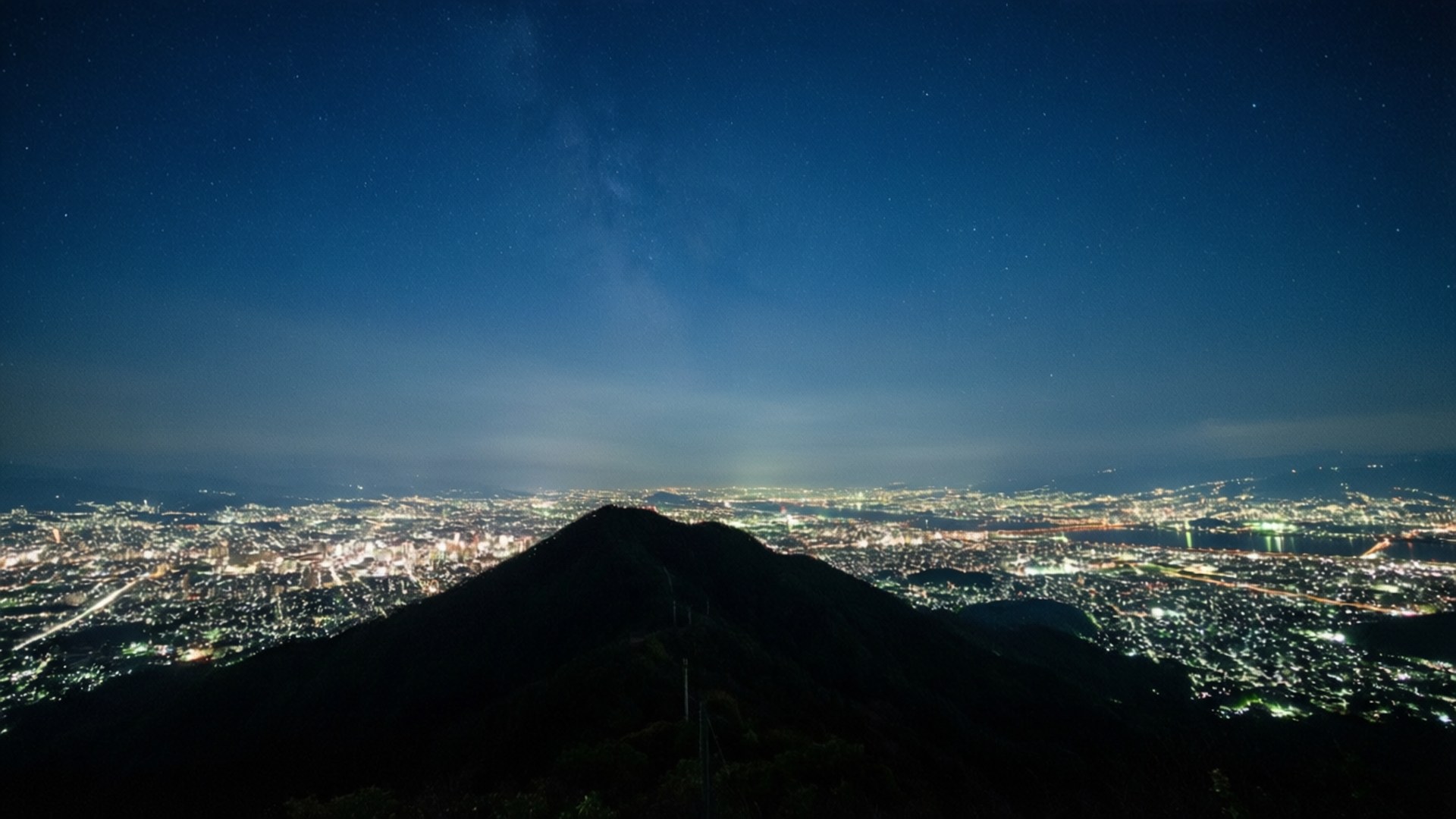 黑暗的山峰俯瞰著夜晚燈火通明的城市，星空下銀河清晰可見