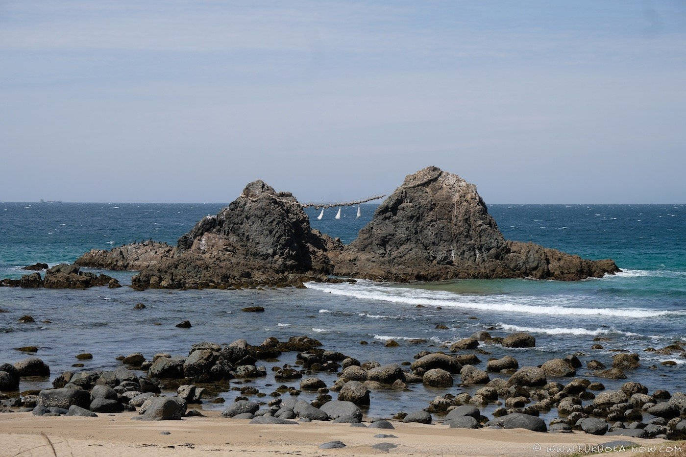 福岡岩石海灘 壁紙 — 一條繩橋連接兩個岩石海蝕柱，上方是淡藍色的天空，附近有藍綠色的海浪拍打。