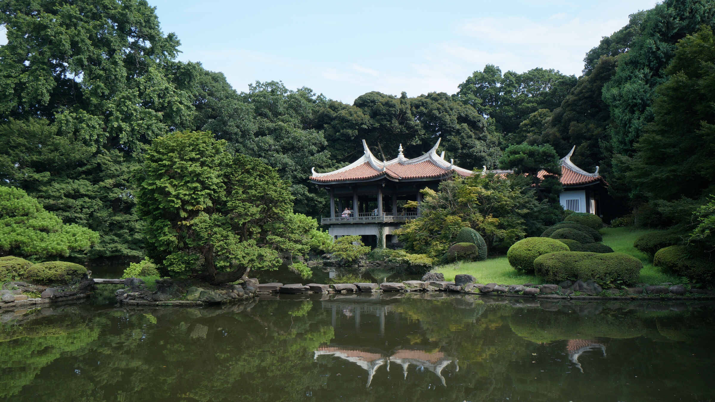 福岡親密森林 壁紙 — 一座傳統東亞亭閣，擁有彎曲的屋頂，寧靜地坐落在平靜的池塘旁，倒映著茂密的綠樹和藍天。