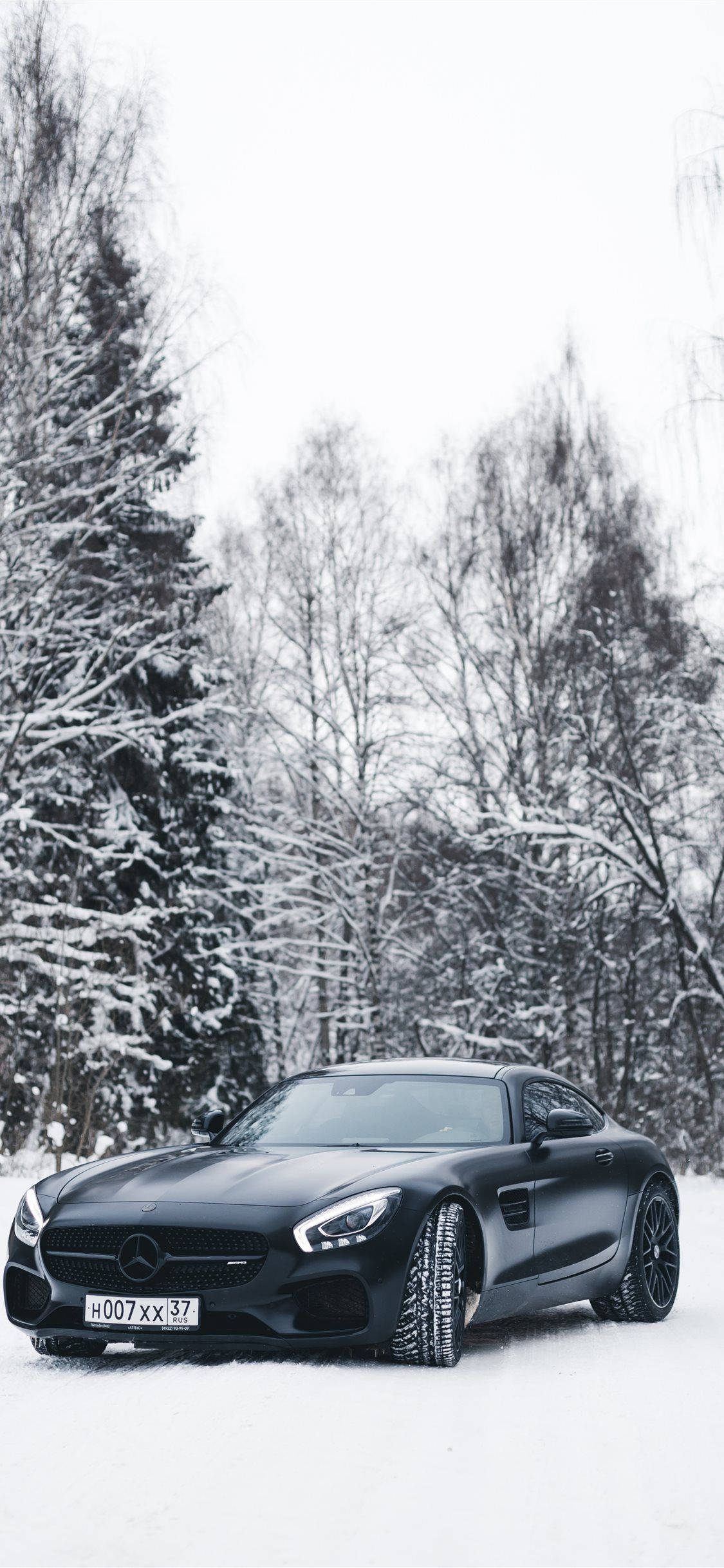 福特 iPhone 雪中駕駛 壁紙 — 一架時尚的啞光黑色跑車停泊在雪地森林中，周圍是白雪覆蓋的樹木，天空多雲。