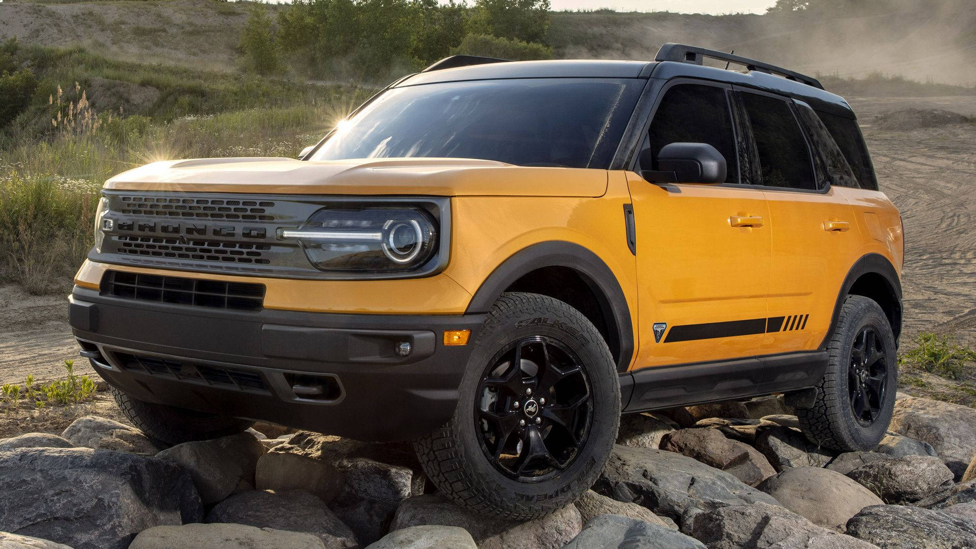 福特Bronco停泊於石堆 壁紙 — 一輛充滿活力的黃色福特Bronco SUV，帶有黑色飾邊和堅固輪胎，在金色陽光下征服岩石地形。
