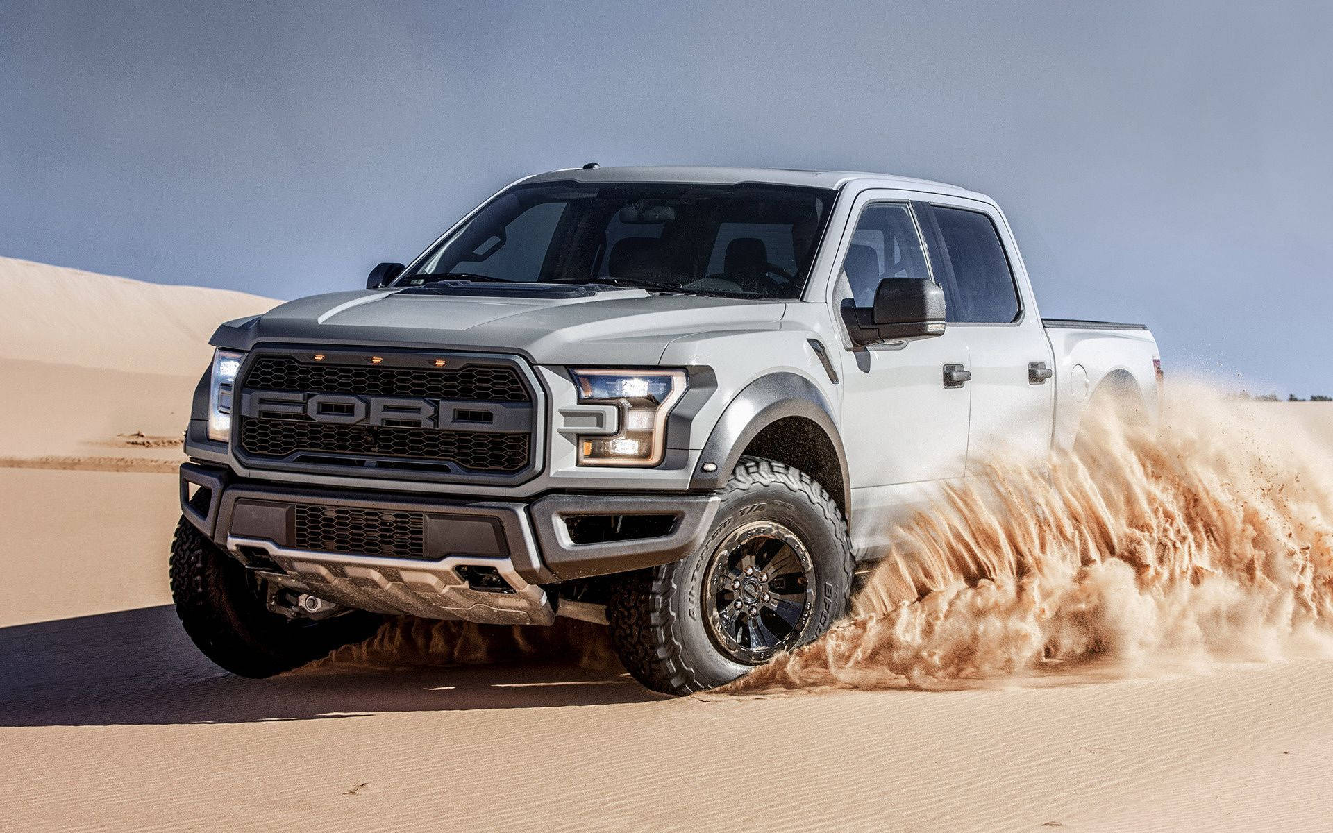 福特Raptor疾馳沙漠 壁紙 — 一輛銀色福特F-150猛禽卡車在晴朗的藍天下穿越沙漠沙丘，揚起巨大的沙塵雲。