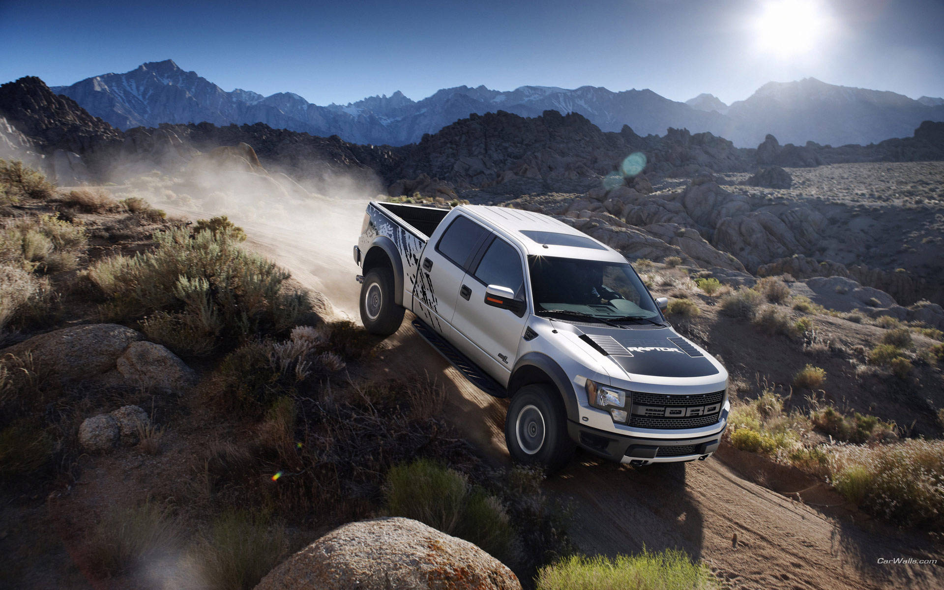 福特Raptor自山巔駛下 壁紙 — 一輛銀色福特 F-150 Raptor 卡車在明亮的陽光下行駛在岩石沙漠小徑上，揚起灰塵，背景是白雪覆蓋的山脈。