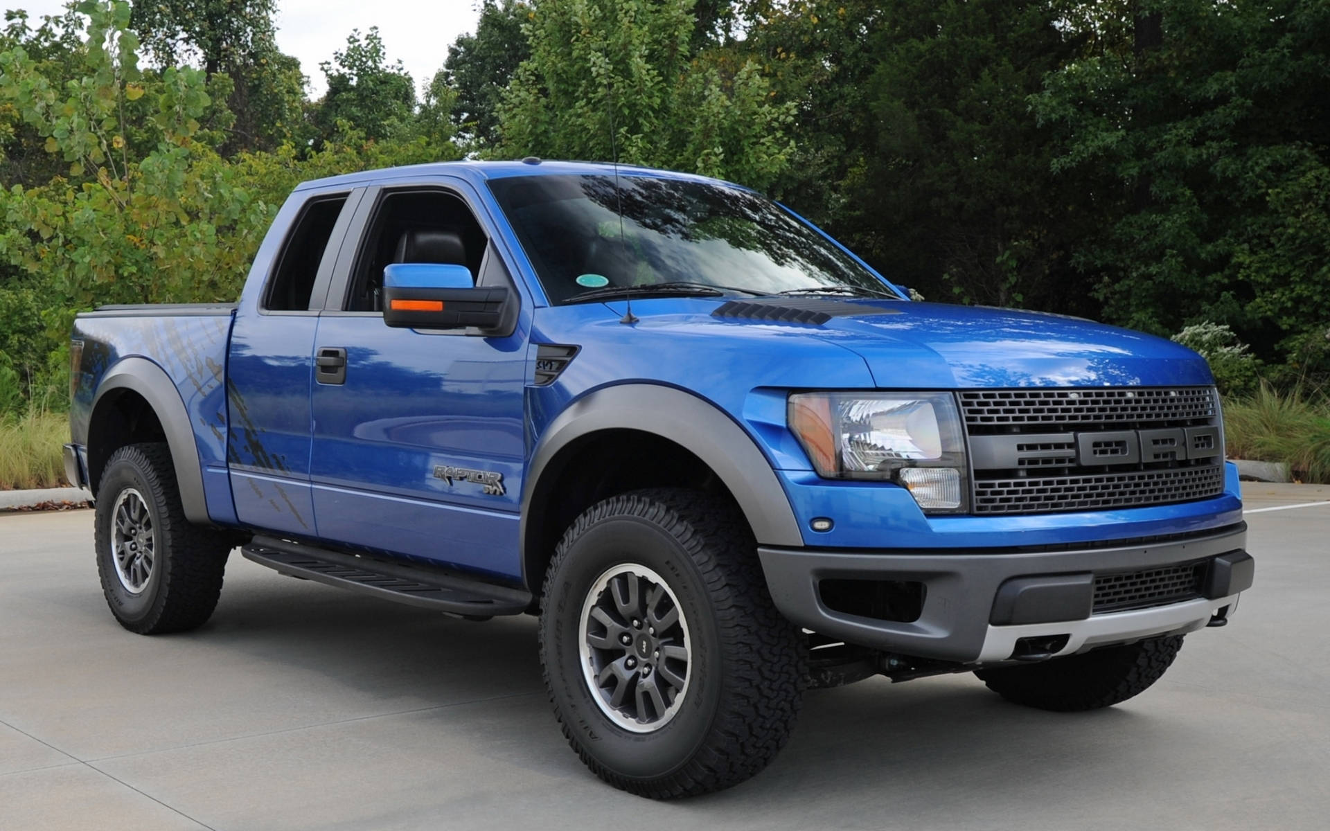 福特Raptor藍光閃電色 壁紙 — 一輛充滿活力的藍色福特 F-150 Raptor 皮卡車停在瀝青路面上，背景是茂密的綠樹。