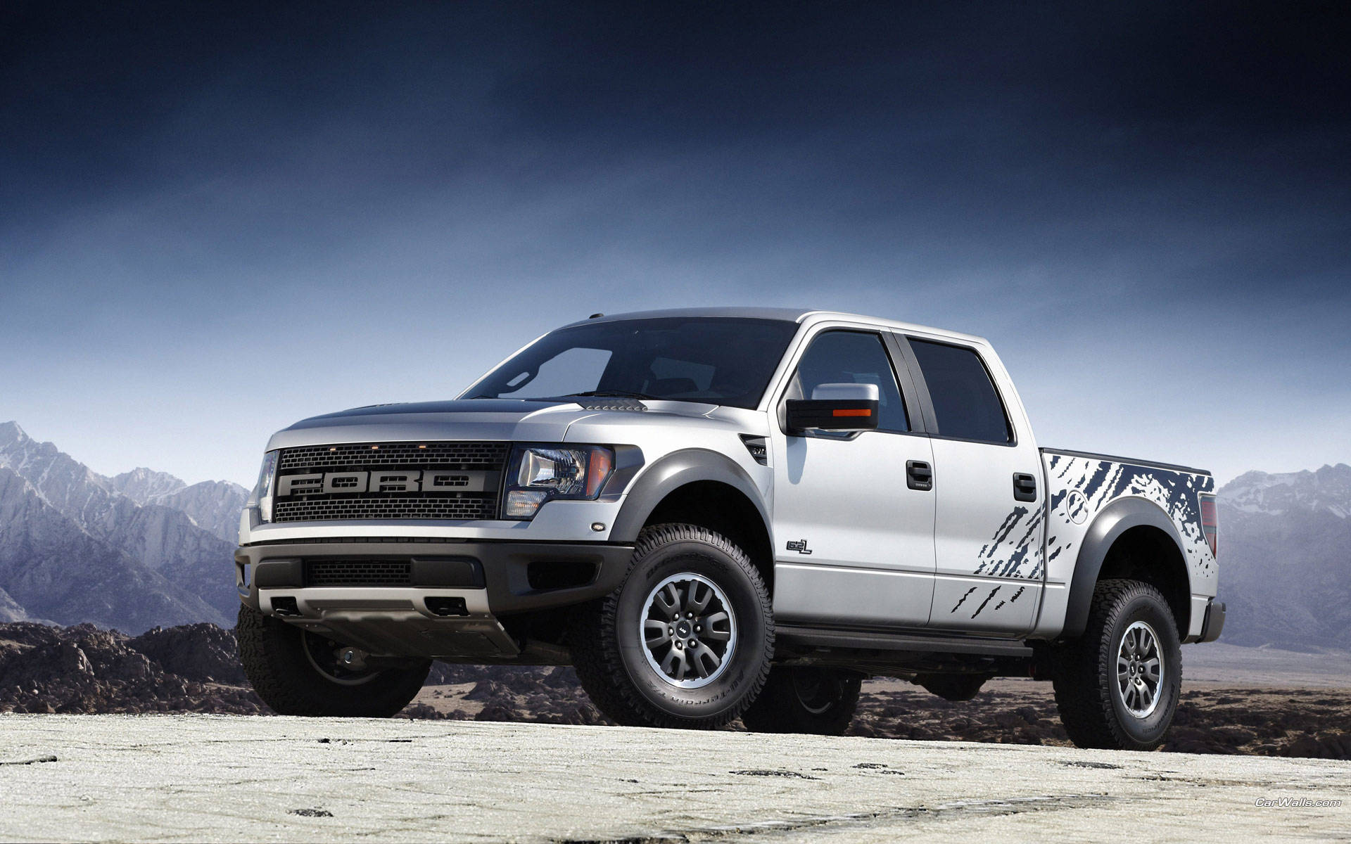 福特Raptor配碳纖維側裙 壁紙 — 一架銀色福特 F-150 Raptor 貨車帶有黑色圖案貼紙，矗立在岩石沙漠平原上，背景是白雪覆蓋的山脈，天空呈現戲劇性的暮色。