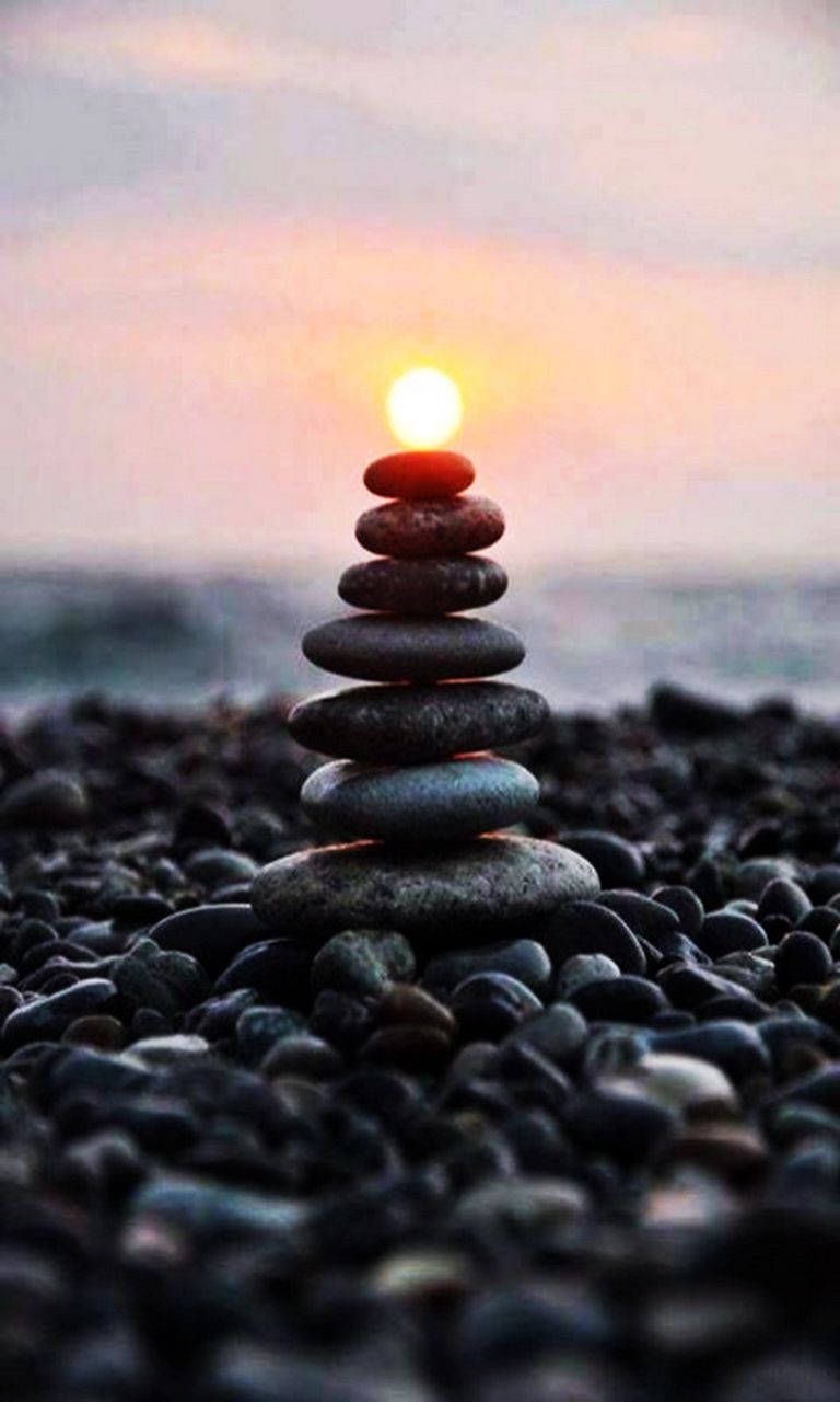 禪黑卵石 壁紙 — 一疊光滑的石頭在卵石海灘上保持平衡，夕陽在後方落下，散發出溫暖的光芒。