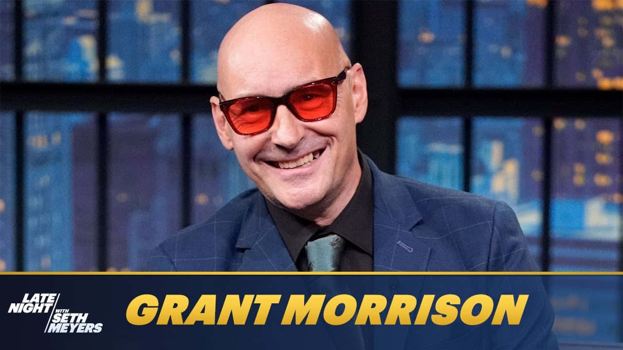 Grant Morrison 桌布 壁紙 — 禿頭男子戴著紅色太陽眼鏡，穿著藍色西裝，在模糊的城市景觀背景下咧嘴大笑。