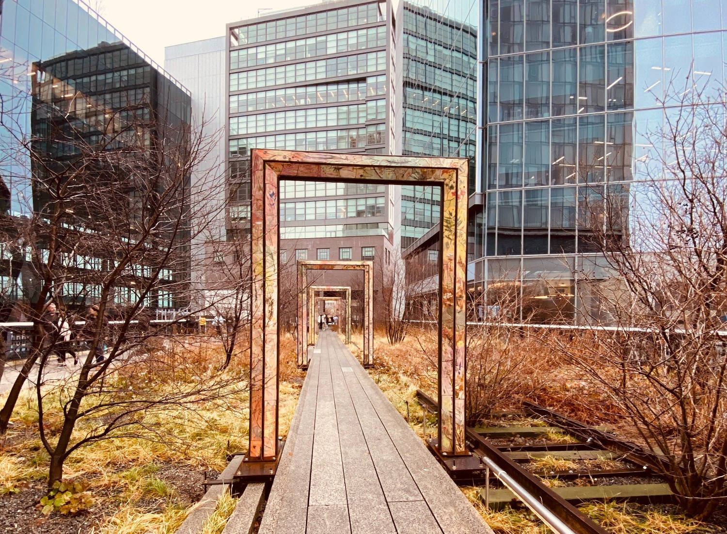 秋季的高線公園 壁紙 — 一條木製步道穿過生鏽的金屬框架，位於被玻璃摩天大樓環繞的草地城市公園中。