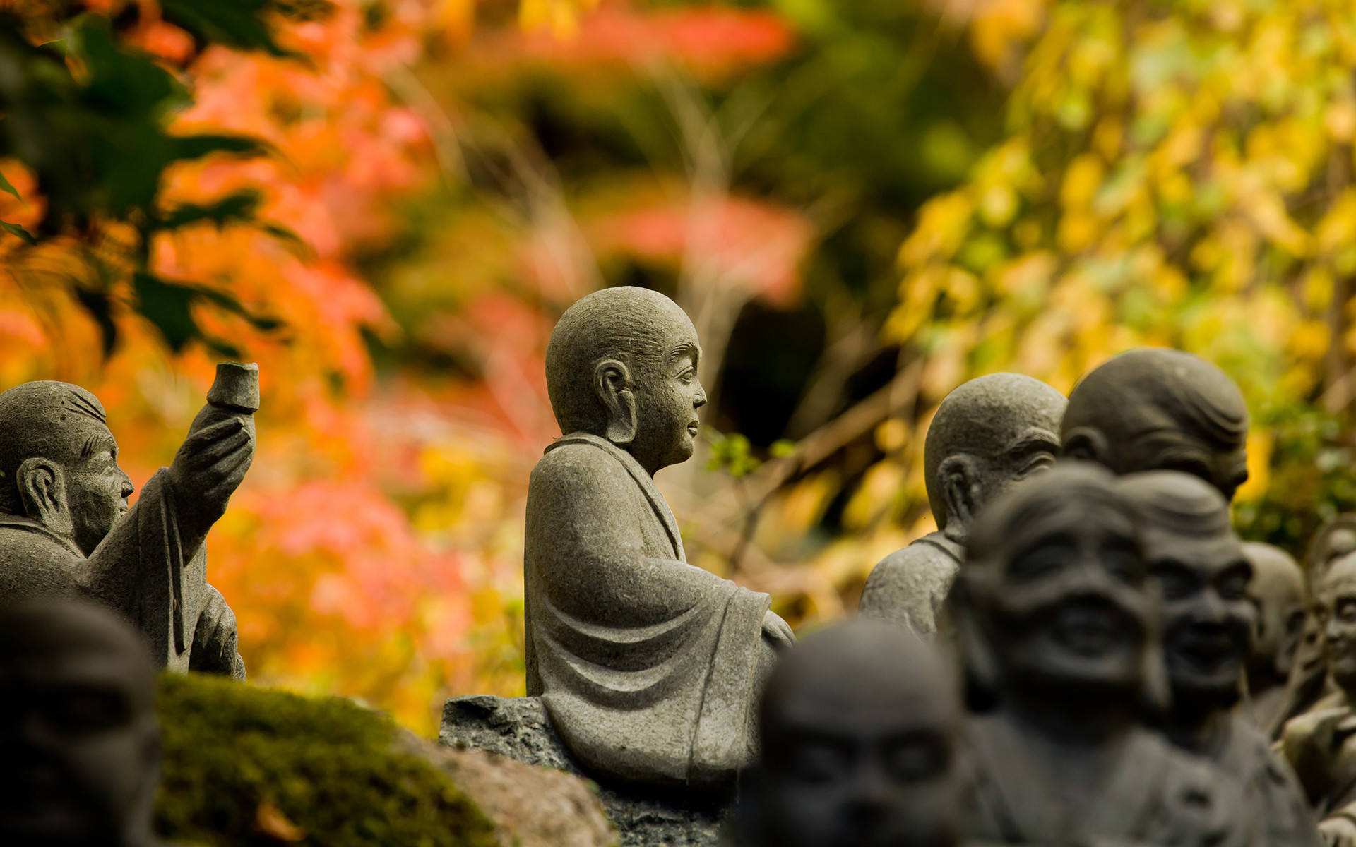 秋日佛像 HD 壁紙 — 寧靜僧侶的石像坐在秋葉中，背景有鮮豔的紅色和金色葉子模糊。