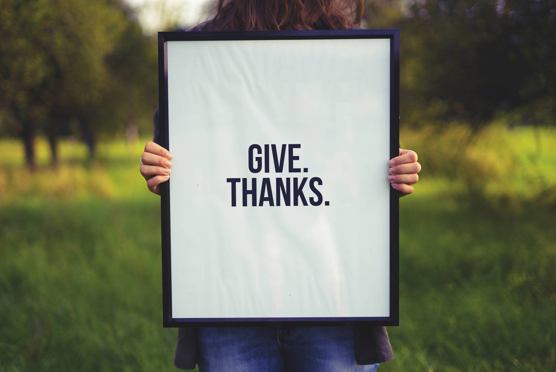 在草地上，一個人拿著一個寫著 give thanks 的帶框牌子。