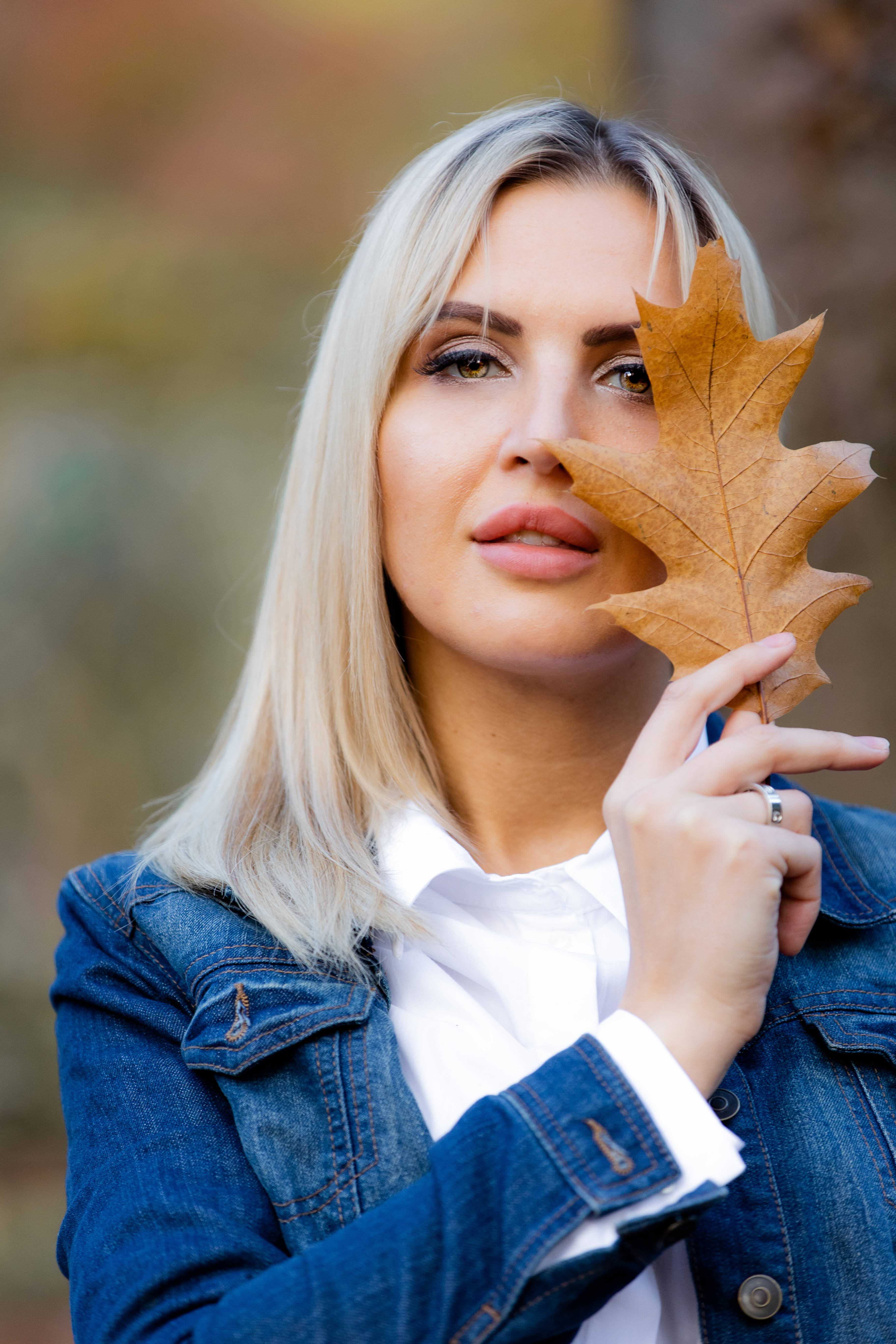 秋日牛仔服少女 壁紙 — 一位金髮女子身穿牛仔外套，手持一片棕色楓葉遮住眼睛，背景是模糊的秋景。