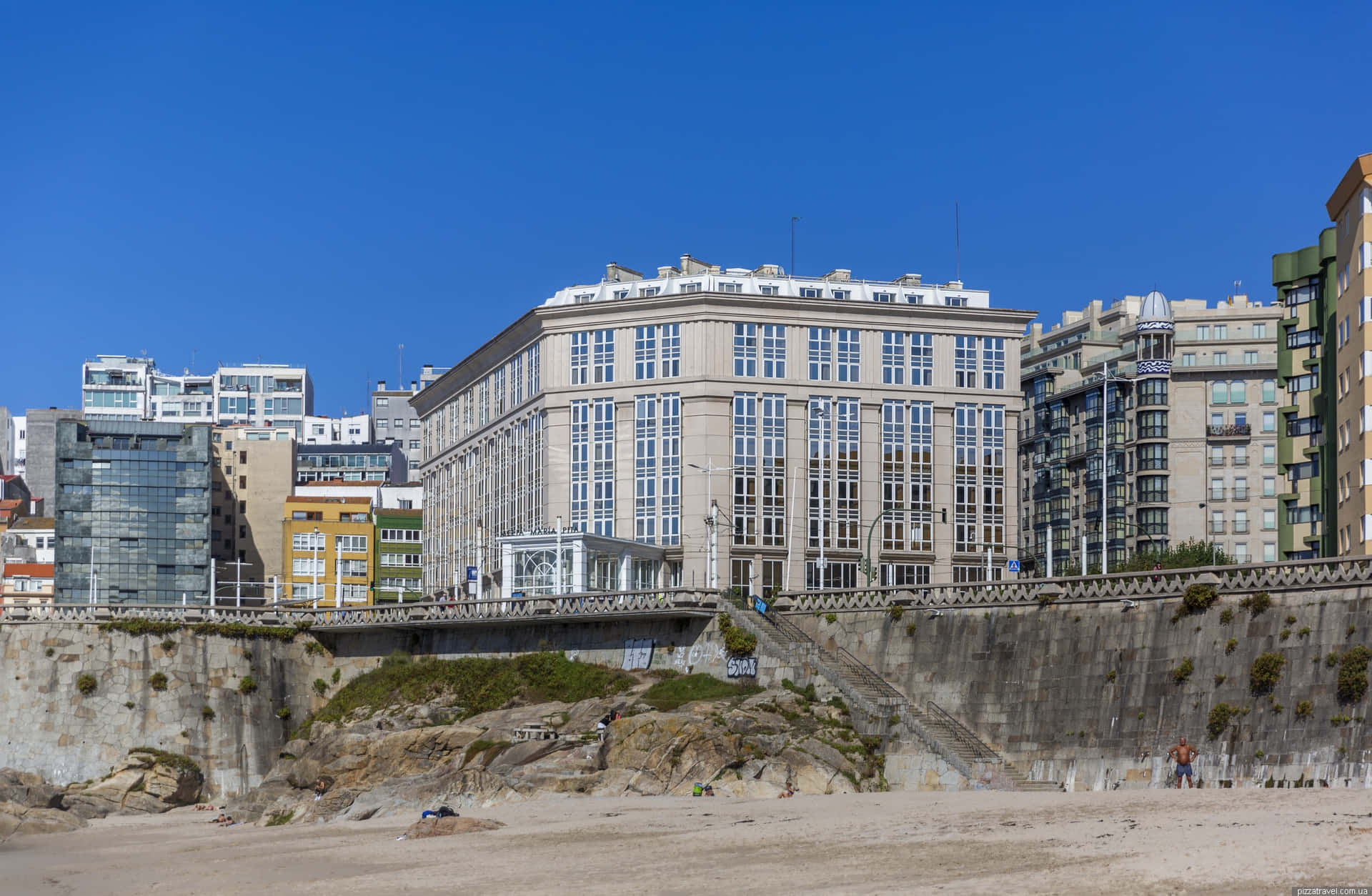 科倫納海灘酒店 壁紙 — 陽光明媚的海濱城市海灘，現代建築在石堤後方聳立。