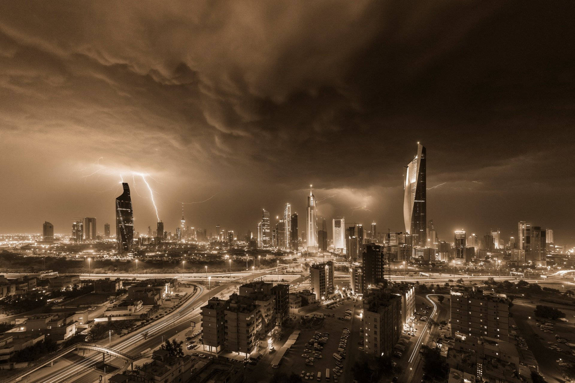 科威特棕褐色風景 壁紙 — 夜晚以深褐色調呈現的戲劇性城市景觀，閃電劃過高聳的摩天大樓，下方街道閃爍著光芒。
