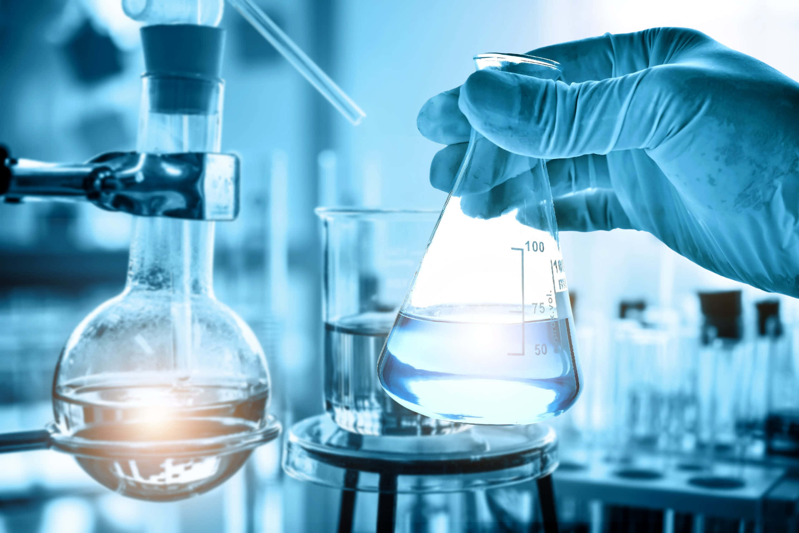 科學實驗室中的突破性發現 壁紙 — 戴手套的手在充滿玻璃器皿的冷藍色實驗室中握住一個發光的燒杯。