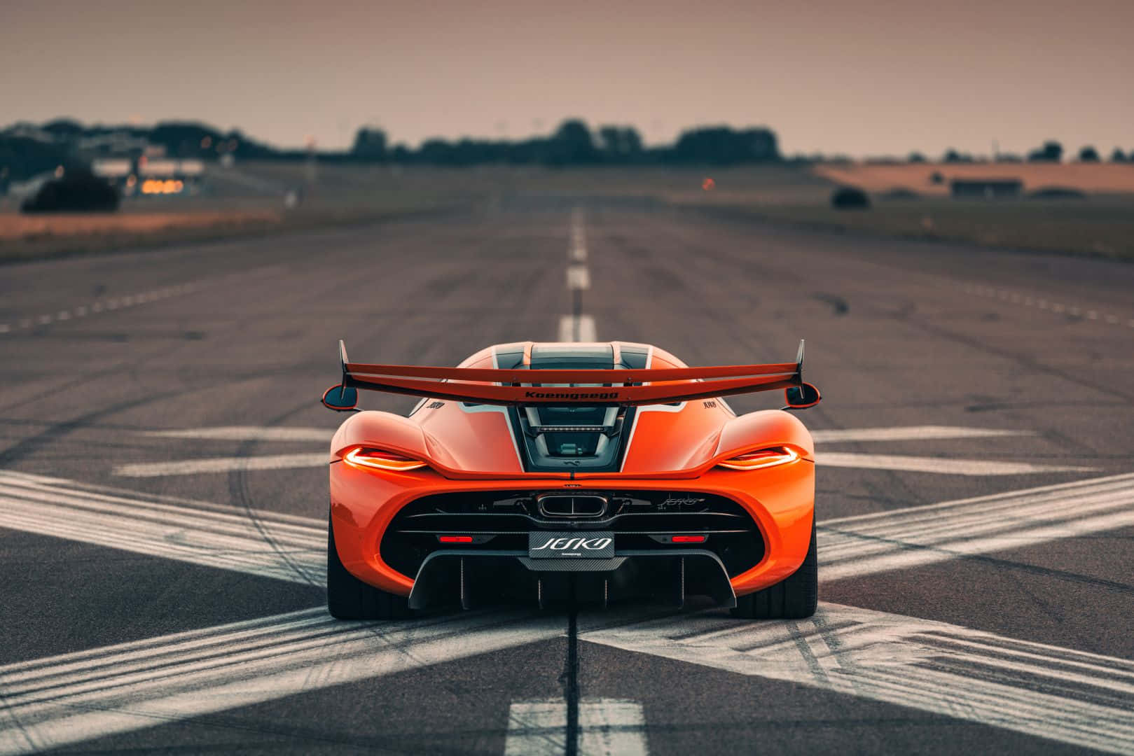 科尼賽格Jesko賽道後視圖 壁紙 — 一輛鮮豔的橙色McLaren Senna跑車在黃昏時分停放在寬闊的瀝青跑道上，尾燈閃爍，天空壯麗。