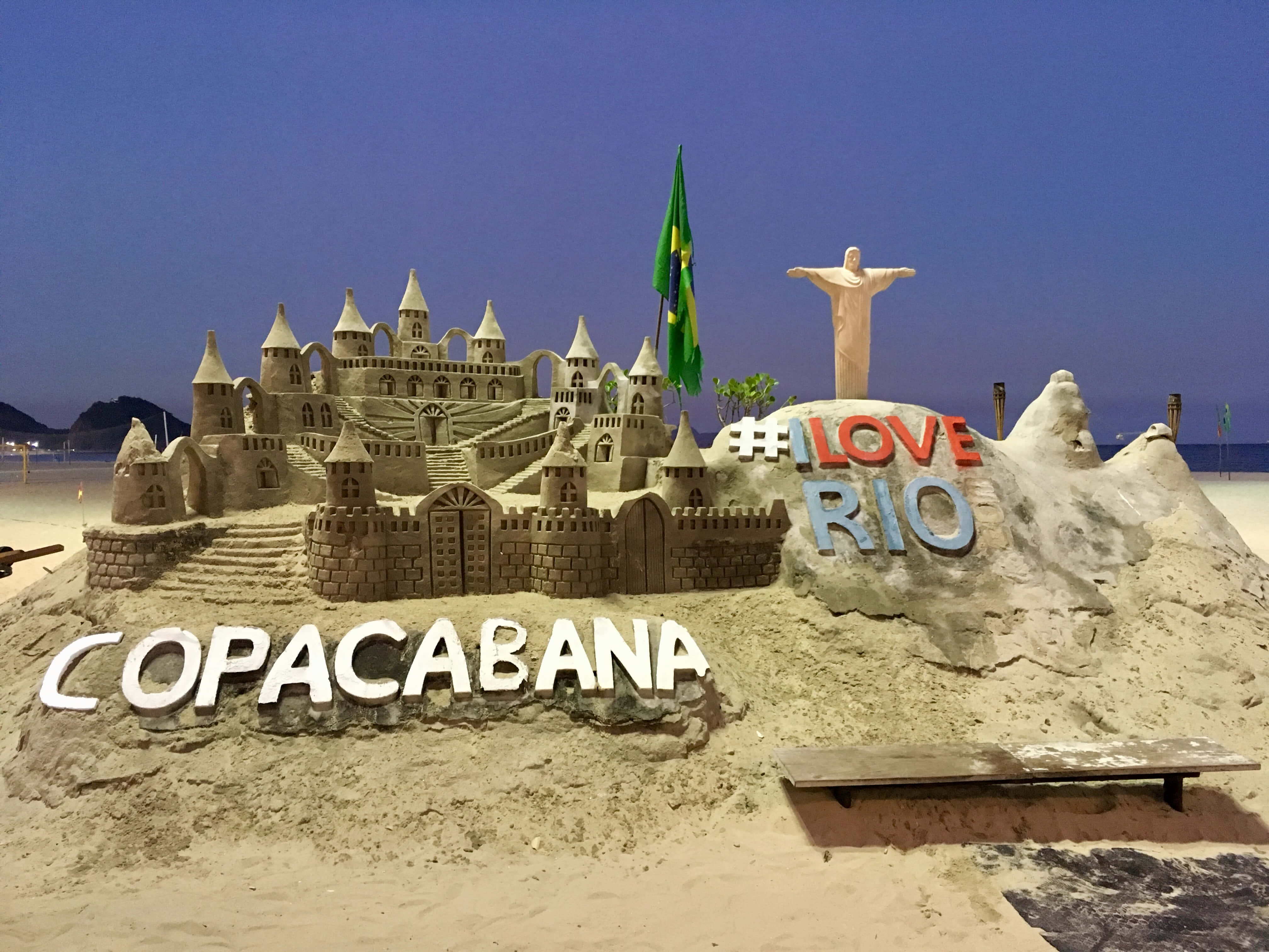 Brazilian Beach 桌布 壁紙 — 科帕卡巴納海灘上的一個巨大沙堡，在暮色天空下展現城堡、基督像和「LOVE RIO」標誌。