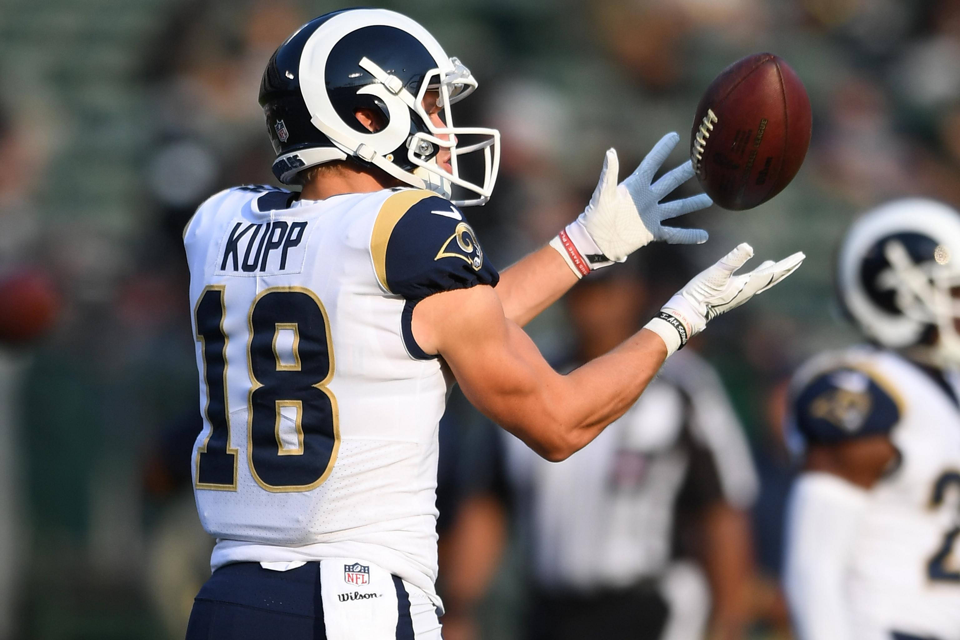 科珀·庫普 NFL 拉姆斯隊美式足球接球攝影 壁紙 — 一位身穿白色和金色球衣的洛杉磯公羊隊美式足球員在比賽中空中接住一個棕色足球。