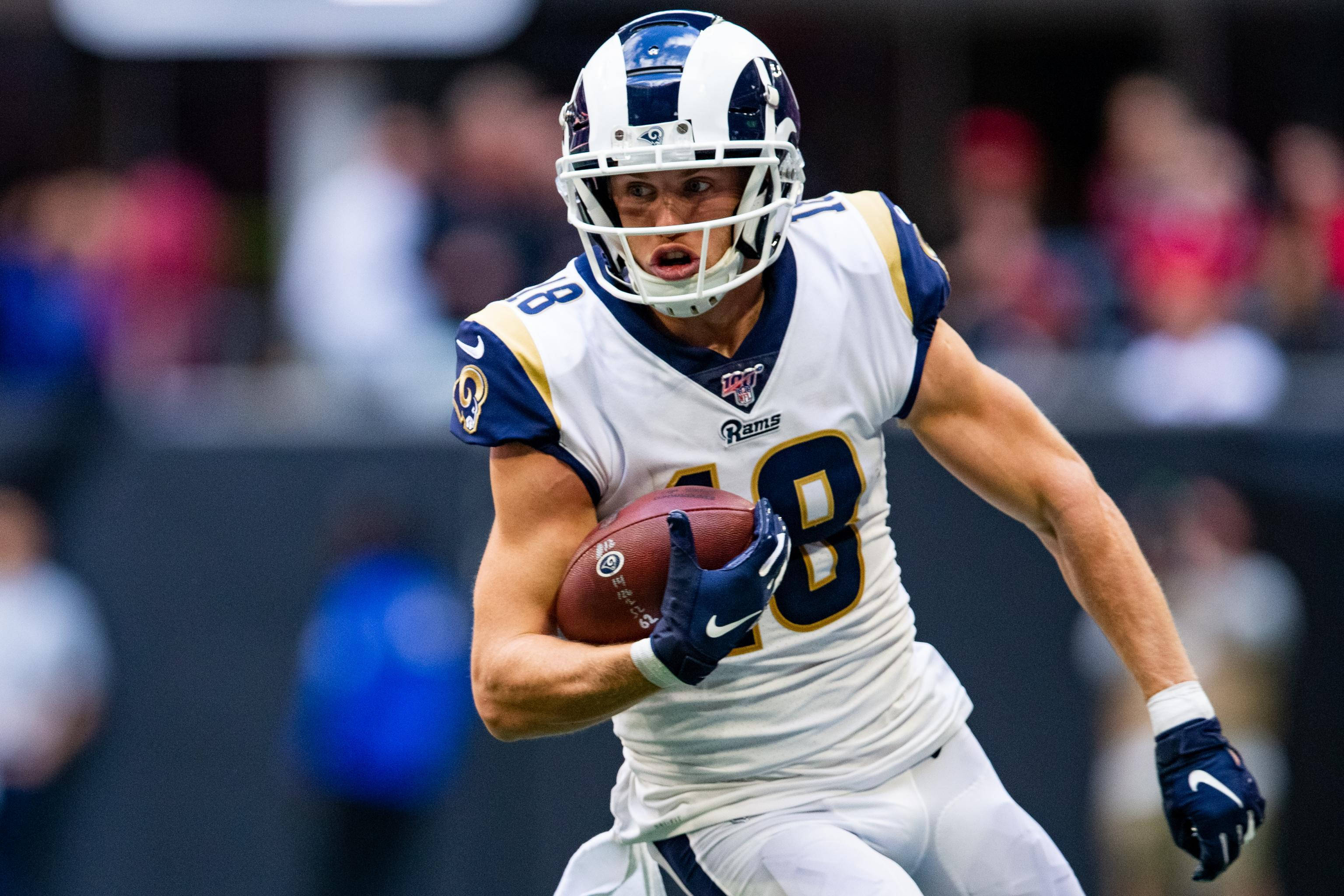 科珀·庫普 NFL 拉姆斯隊美式足球衝鋒攝影 壁紙 — 一名身穿白色和藍色球衣的美式足球員在比賽中向前衝刺，緊握著一個棕色足球。