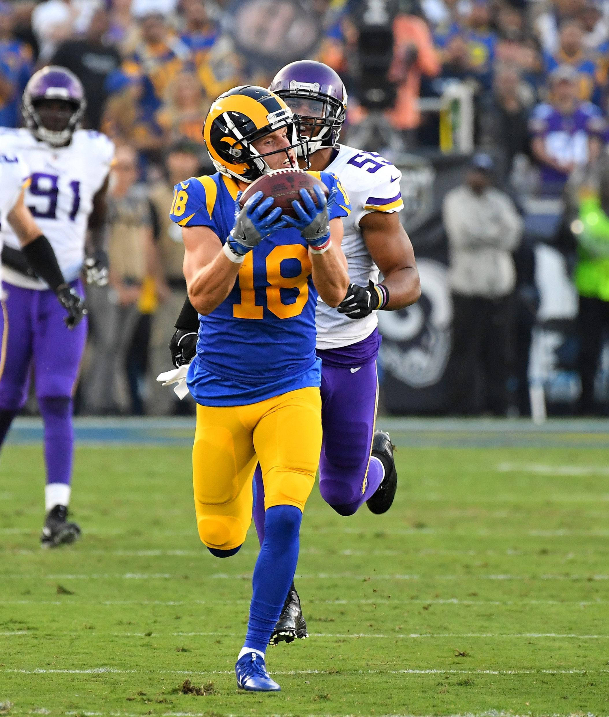 科珀·庫普 NFL 洛杉磯拉姆斯隊跑動接球攝影 壁紙 — 一位身穿藍色和金色18號球衣的洛杉磯公羊隊美式足球員，在充滿活力的綠色球場上接球，同時被明尼蘇達維京人隊的防守球員追趕。