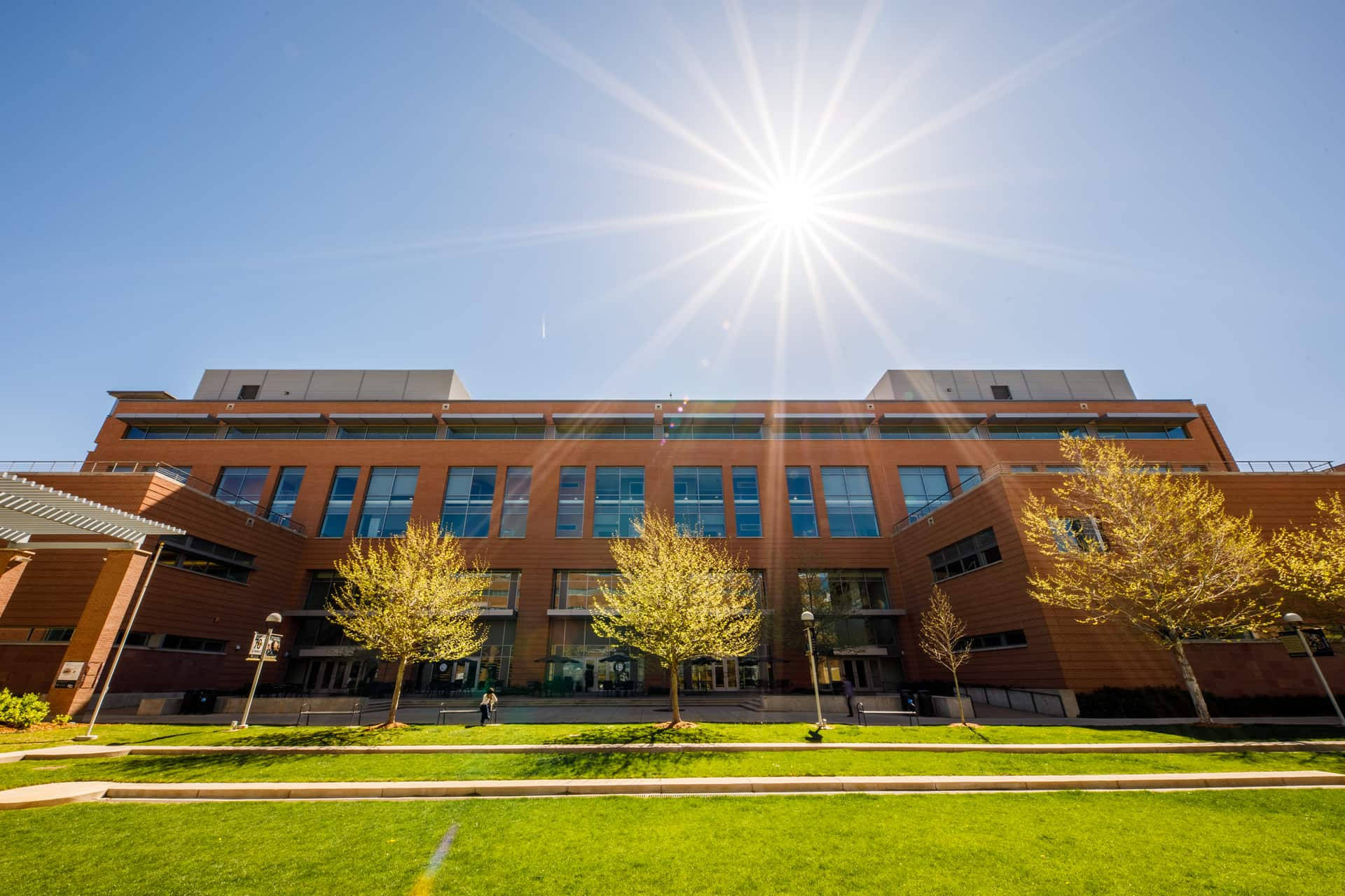 科羅拉多大學校園壯麗鳥瞰 壁紙 — 一座現代磚砌建築，配有玻璃窗，矗立在明亮的陽光下，周圍環繞著金色的樹木和綠色的草坪。
